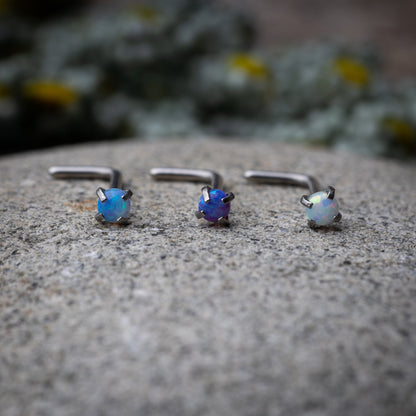 Three L-shaped opalescent nose studs in different shades, displayed on a stone background.
