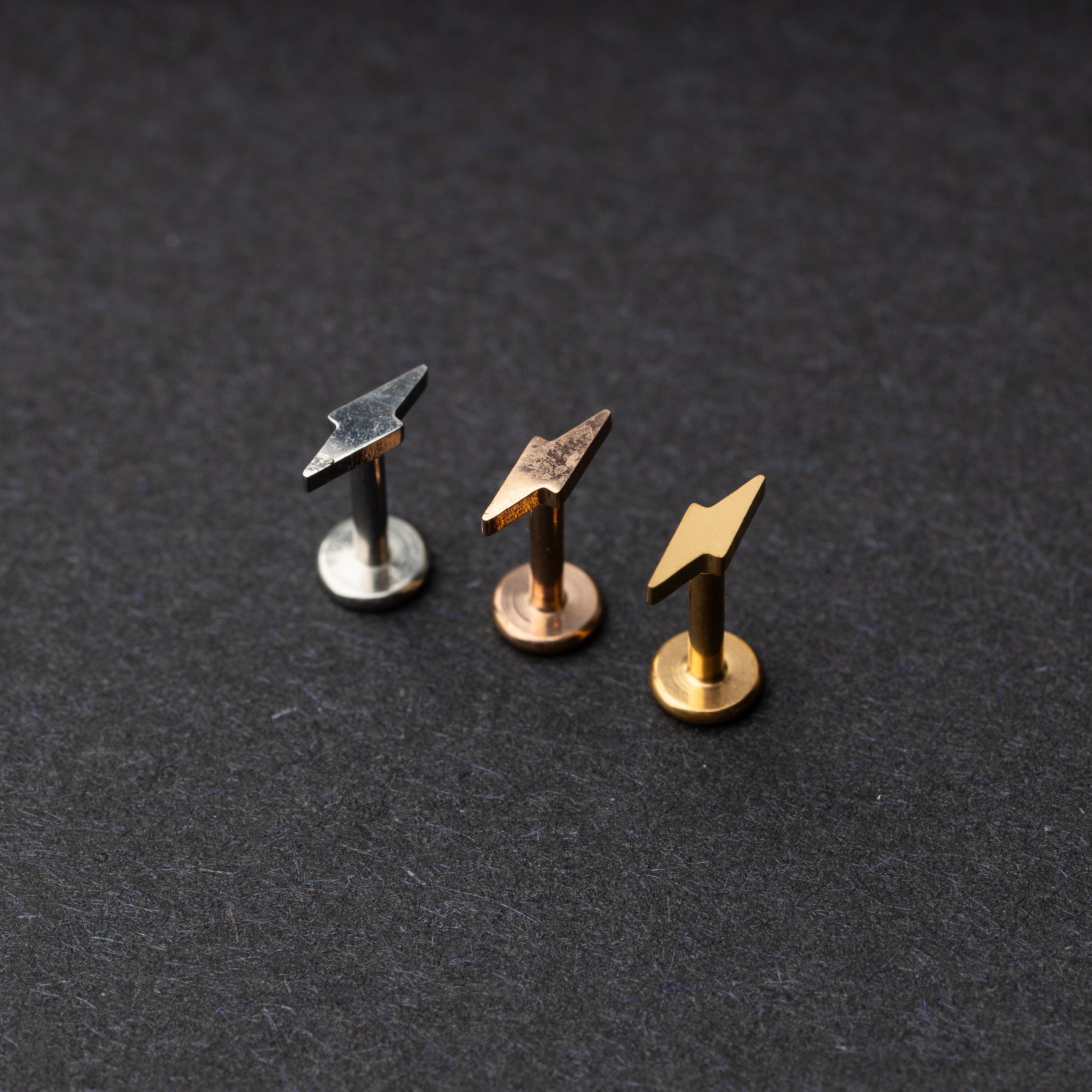 Minimalist lightning bolt labret studs in silver, gold, and rose gold finishes, displayed on a dark background.
