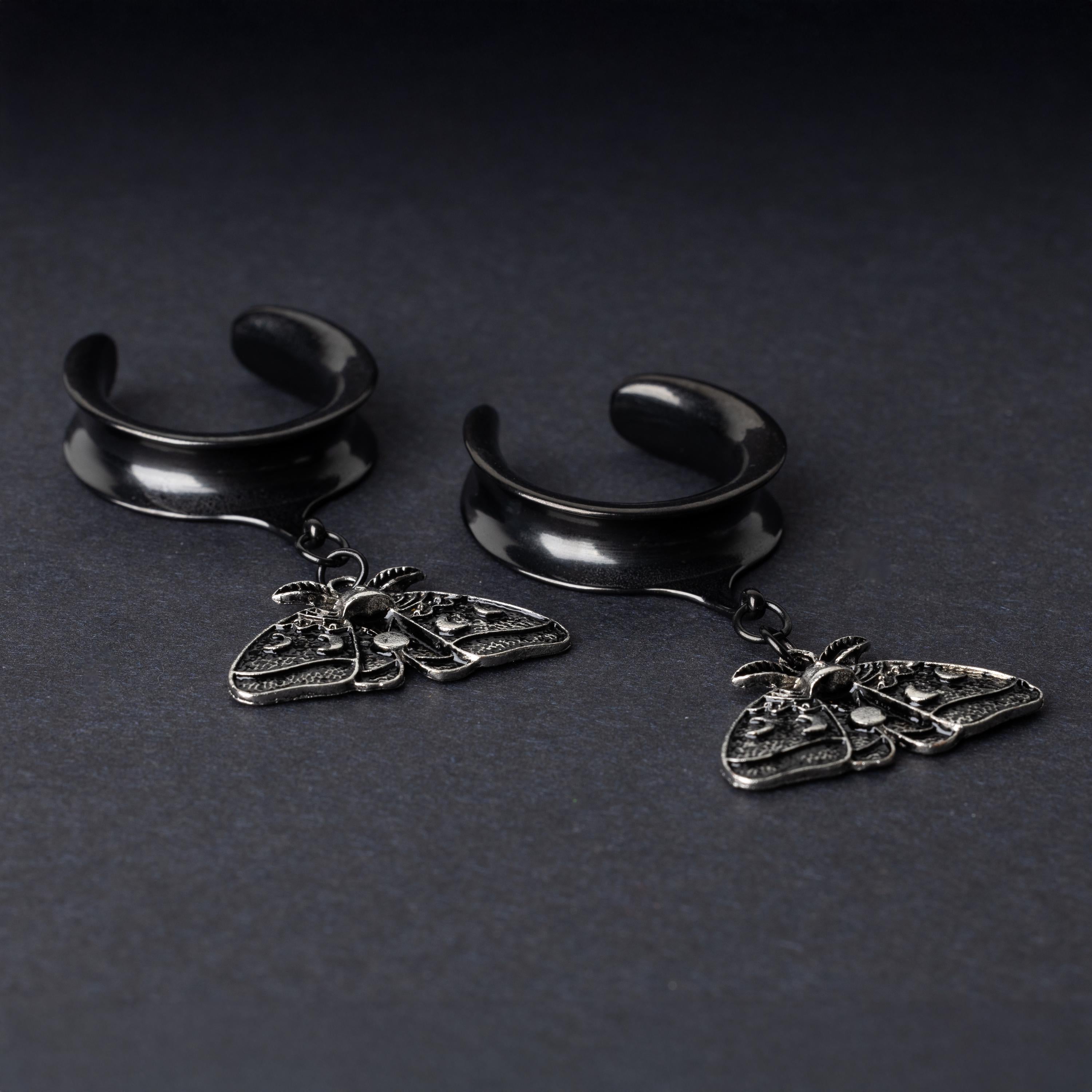 Black tunnels for stretched ears with dangling ornate moth charms featuring celestial patterns, displayed on a dark background.