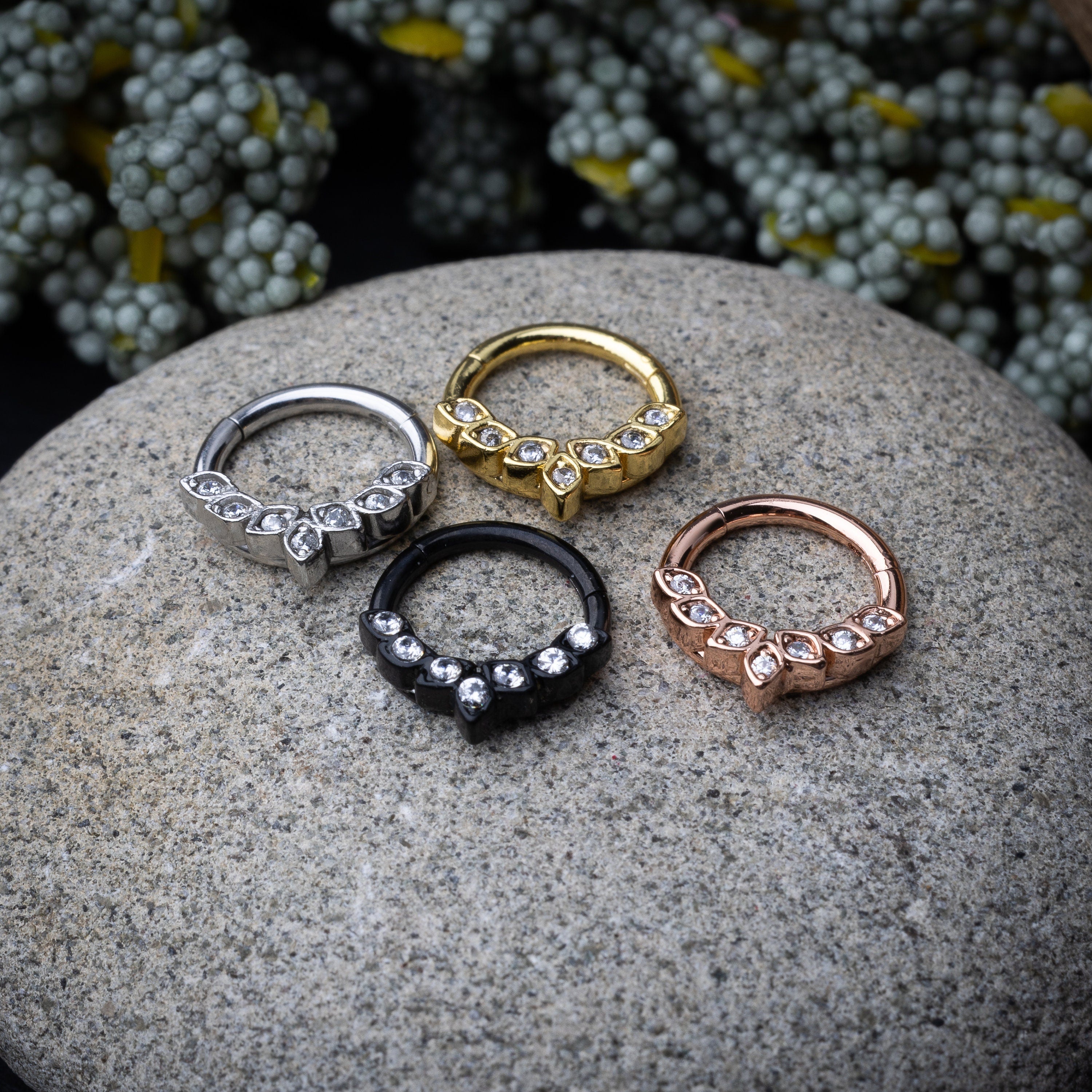 Four marquise gemstone stainless steel clickers in rose gold, gold, silver, and black finishes, arranged on a stone background.
