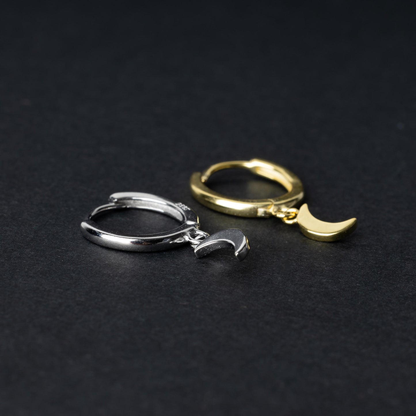 Two crescent moon huggie hoop earrings in gold and silver, each featuring a dangling crescent charm, displayed on a dark background.