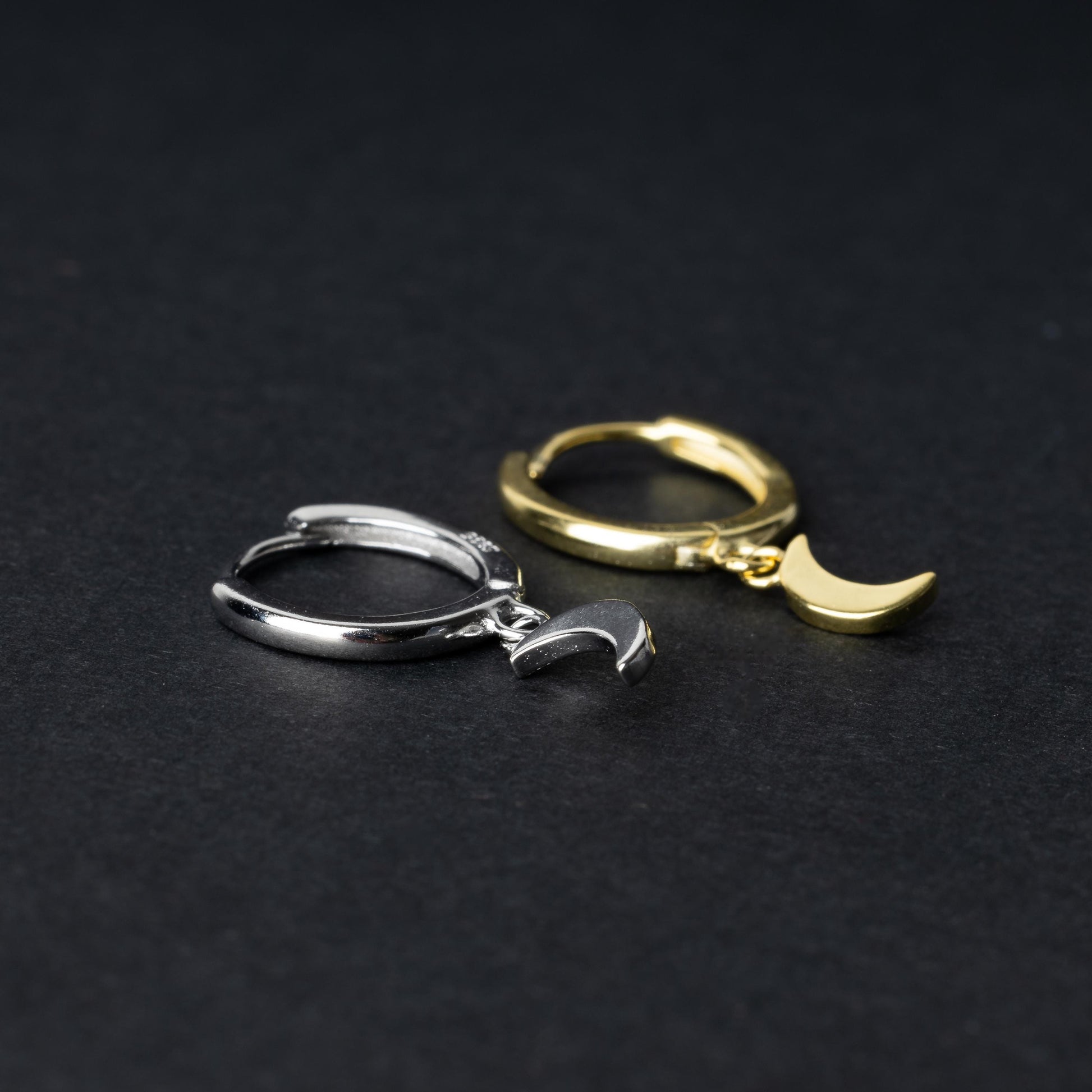 Two crescent moon huggie hoop earrings in gold and silver, each featuring a dangling crescent charm, displayed on a dark background.