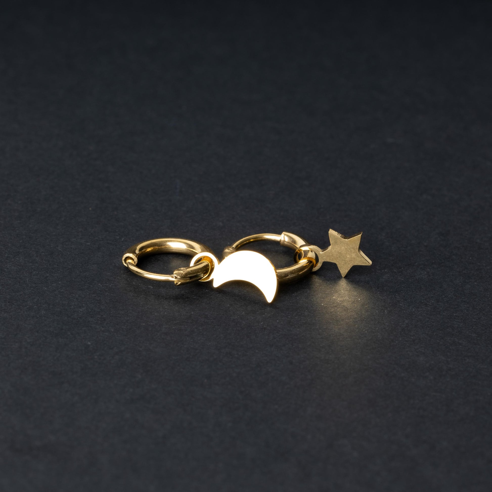 Gold huggie hoop earrings featuring a crescent moon charm on one hoop and a star charm on the other, displayed on a dark background.