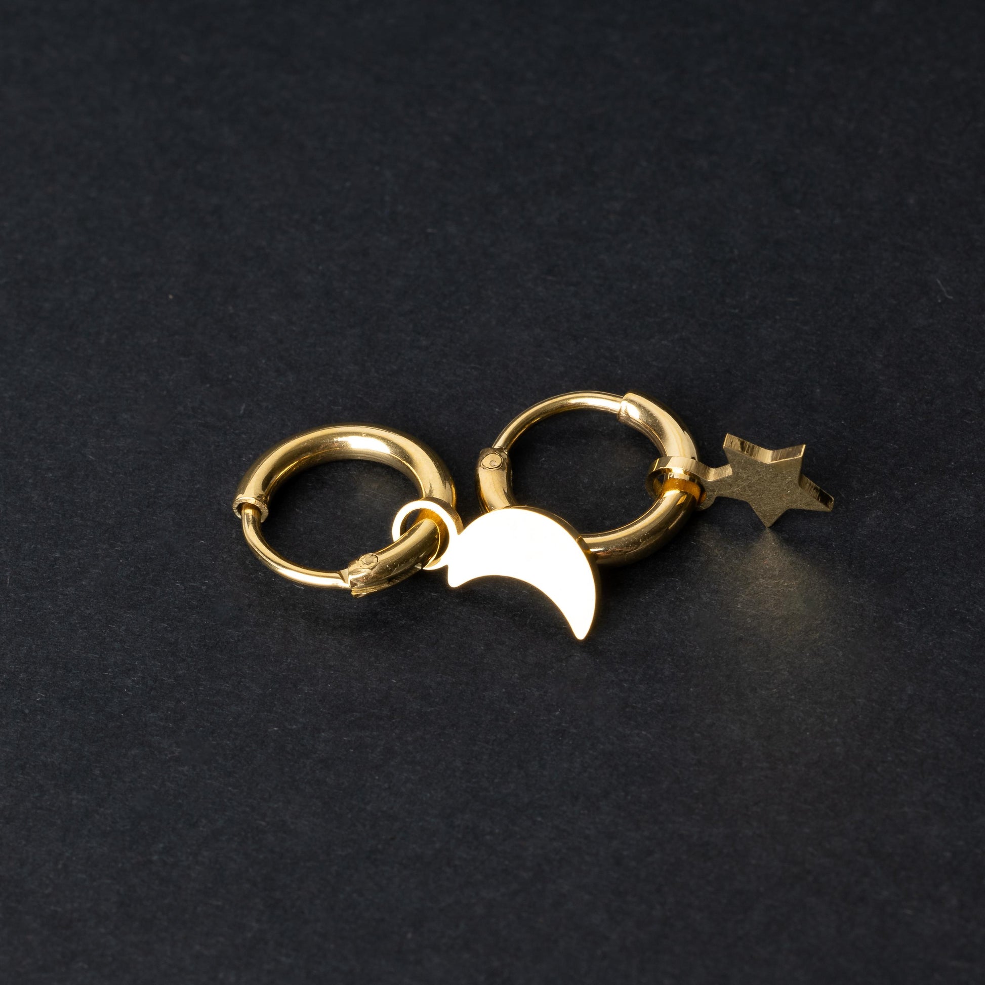 Gold huggie hoop earrings featuring a crescent moon charm on one hoop and a star charm on the other, displayed on a dark background.