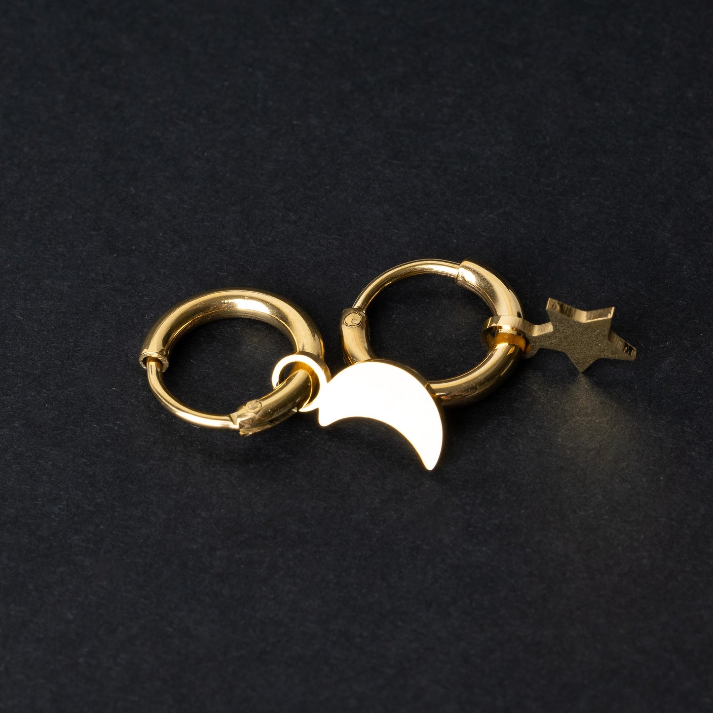 Gold huggie hoop earrings featuring a crescent moon charm on one hoop and a star charm on the other, displayed on a dark background.
