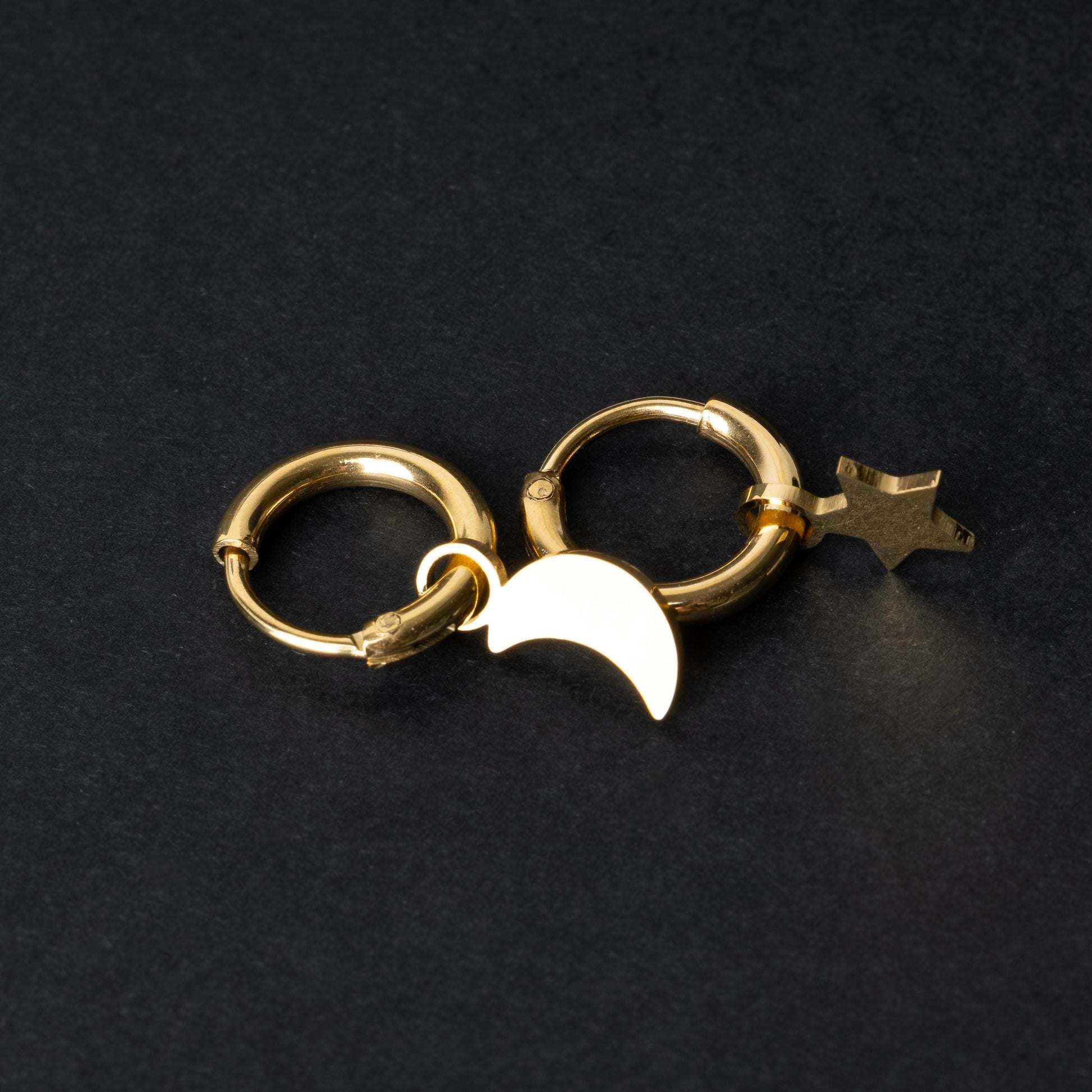 Gold huggie hoop earrings featuring a crescent moon charm on one hoop and a star charm on the other, displayed on a dark background.