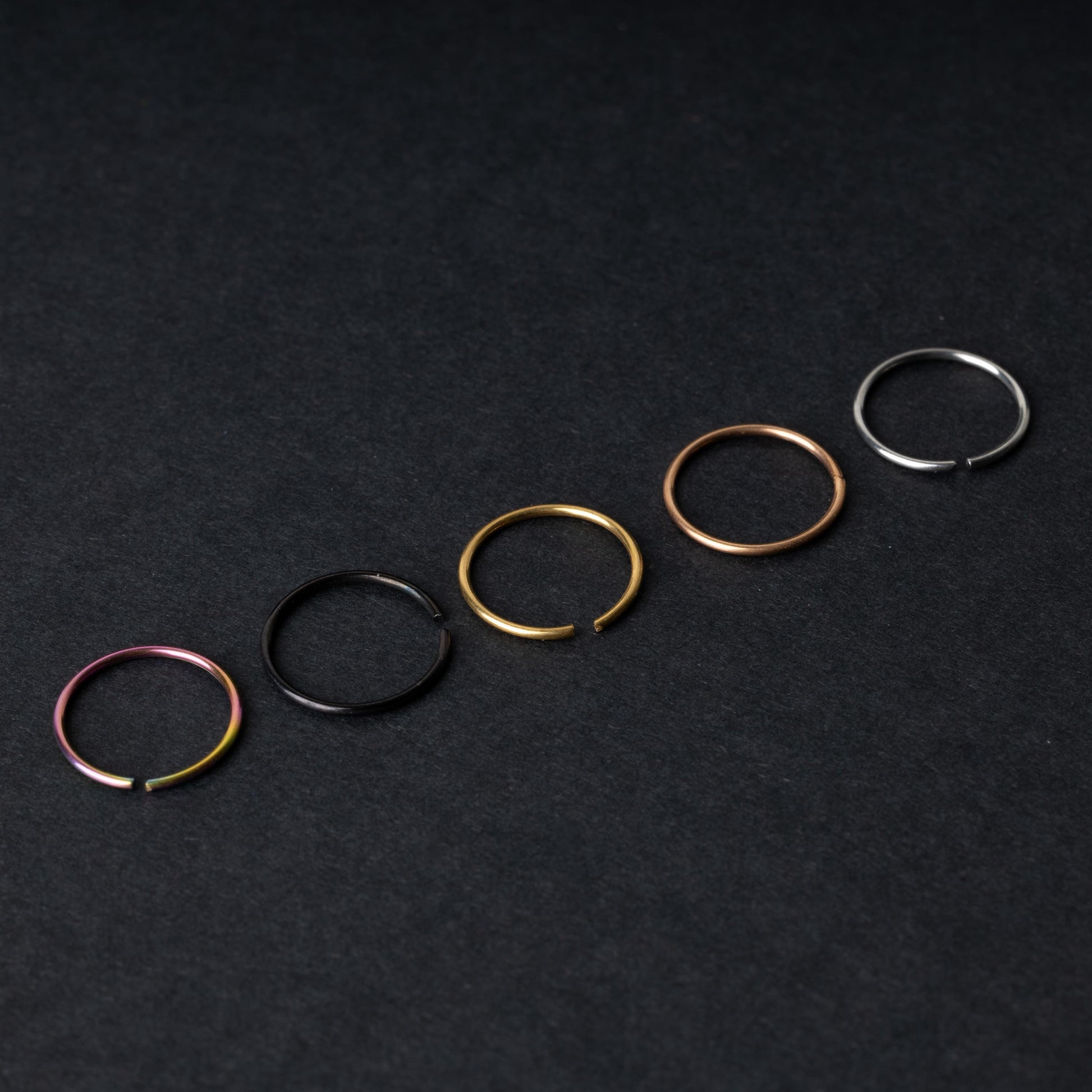 Five thin seamless hoops in rainbow, black, gold, rose gold, and silver finishes, displayed on a dark background.