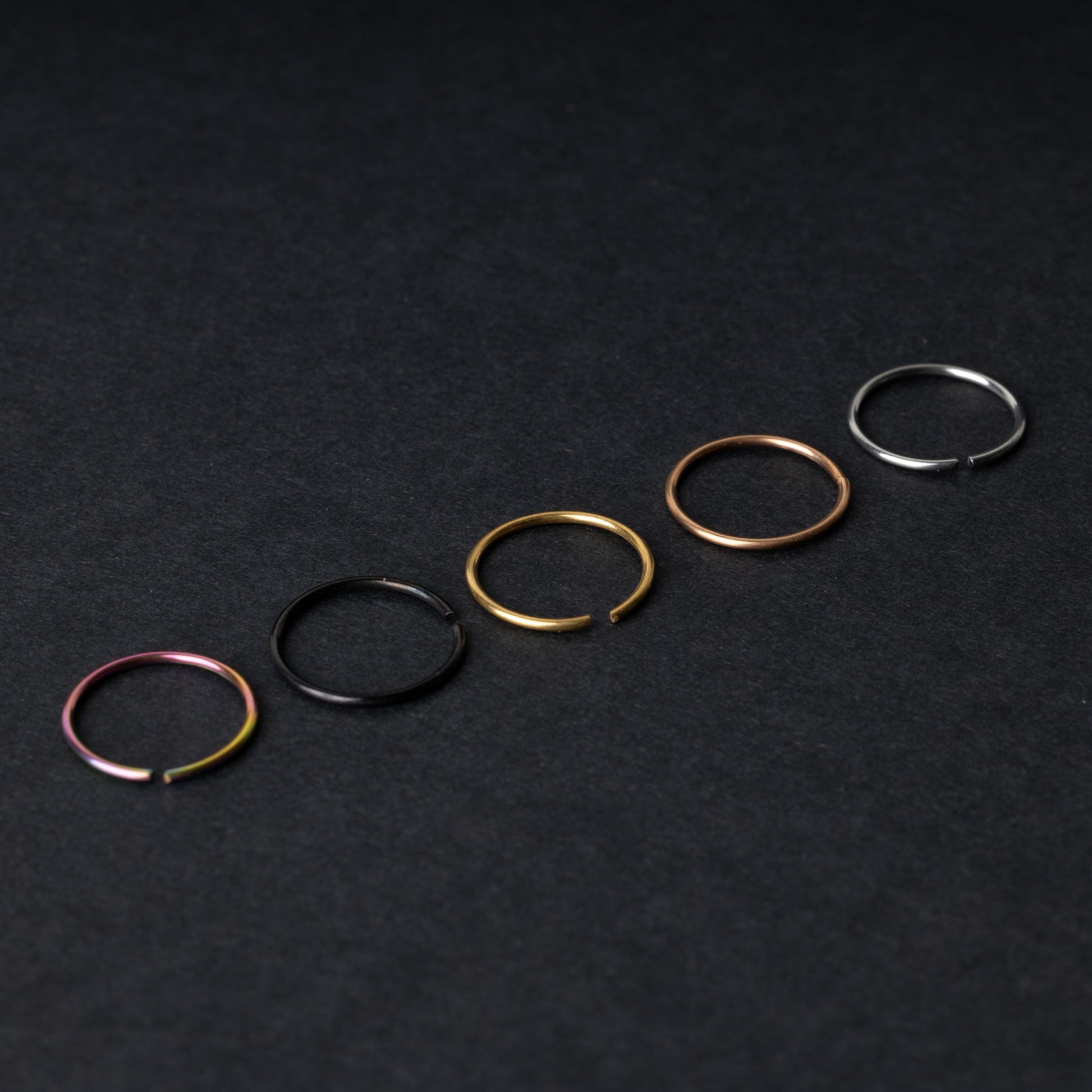 Five thin seamless hoops in rainbow, black, gold, rose gold, and silver finishes, displayed on a dark background.
