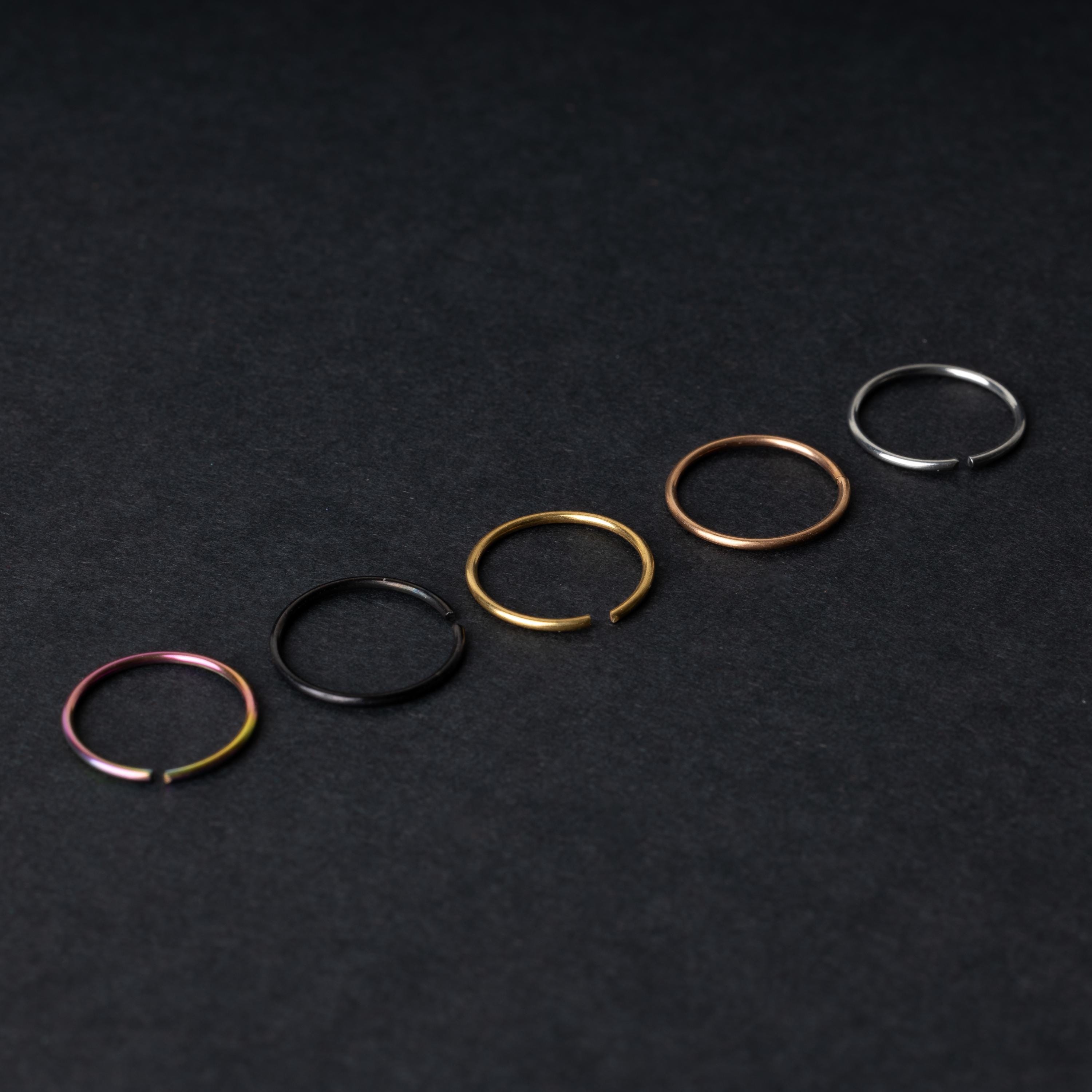 Five thin seamless hoops in rainbow, black, gold, rose gold, and silver finishes, displayed on a dark background.