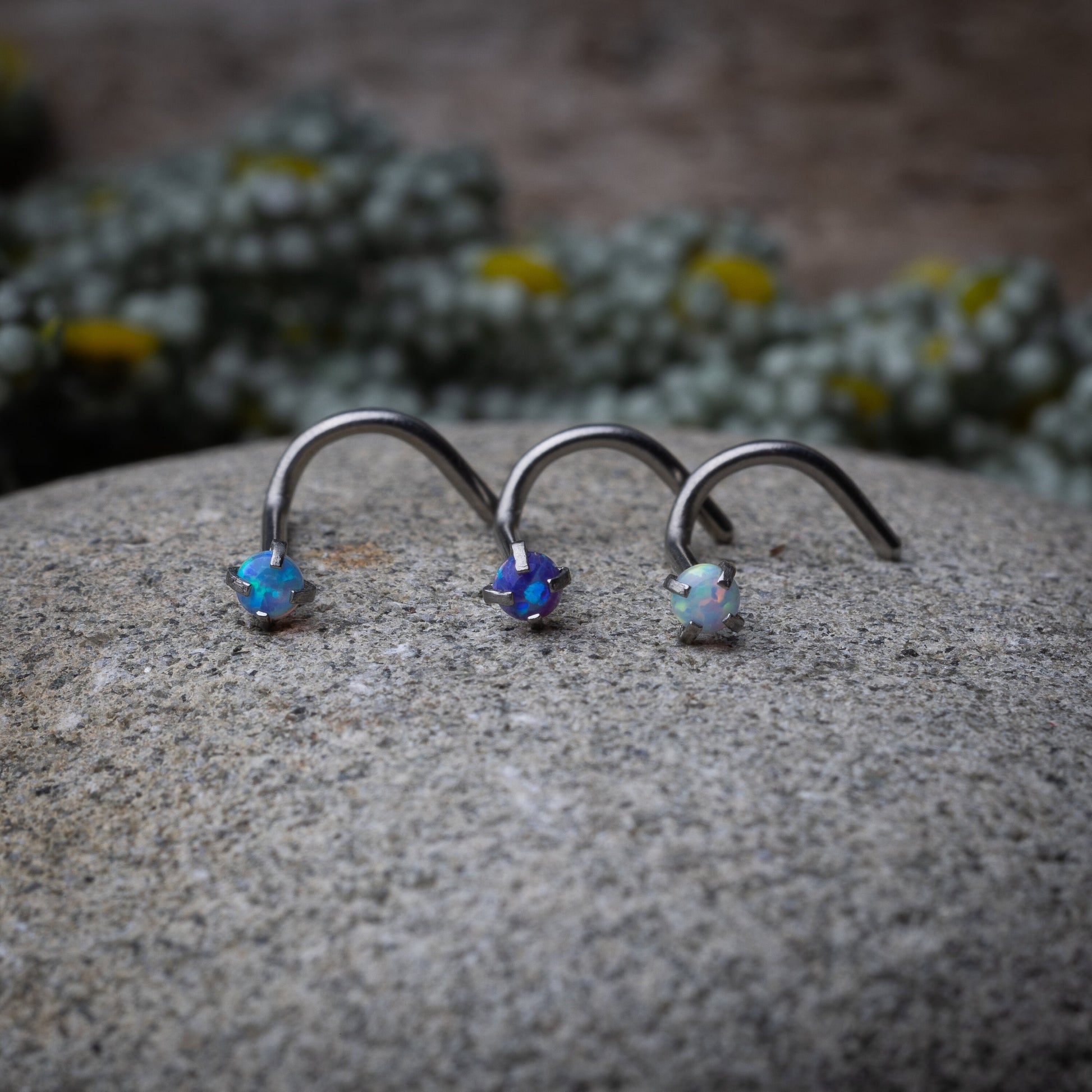 Opalescent nose screws in various colors with a prong setting, displayed on a stone background.