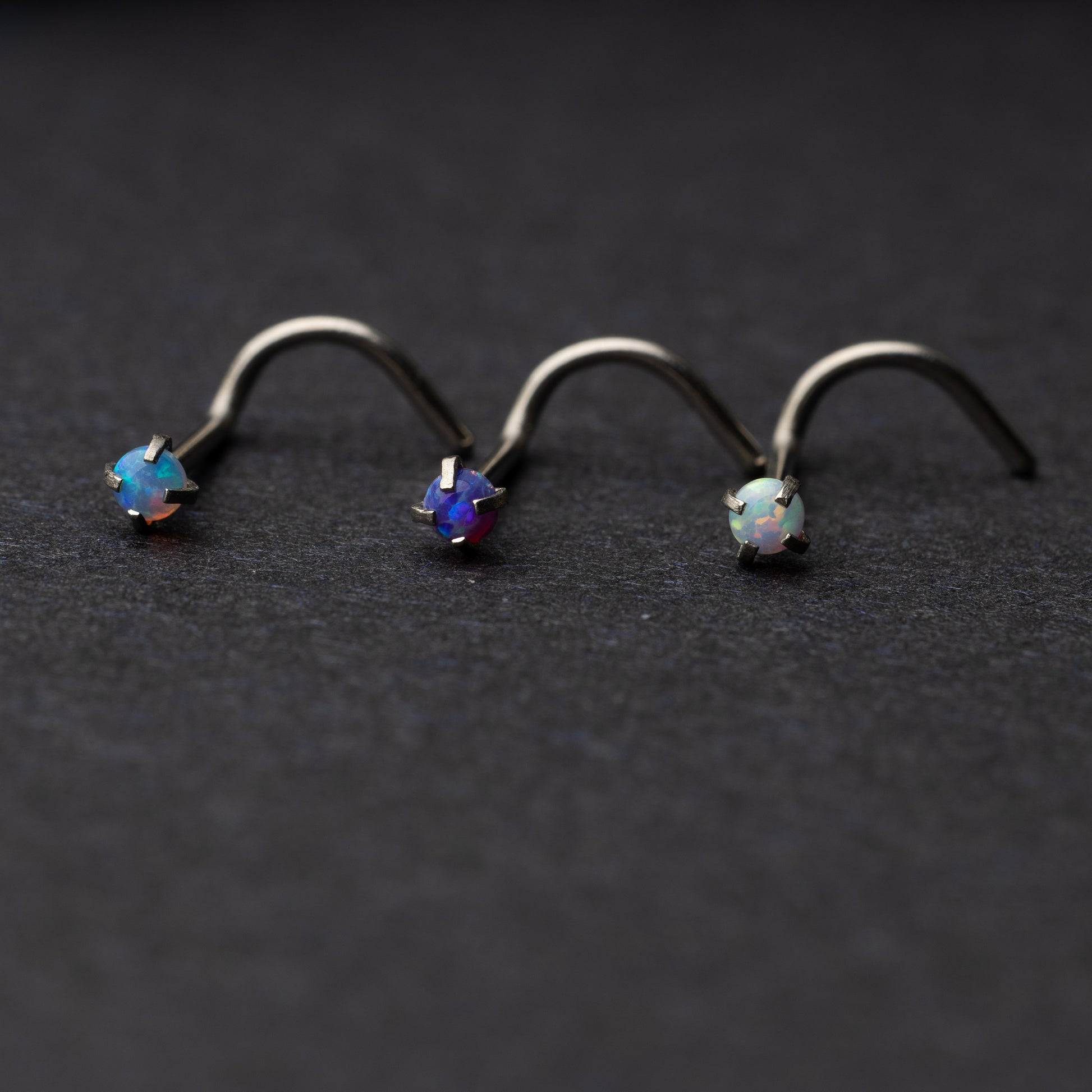 Opalescent nose screws in various colors with a prong setting, displayed on a dark background.