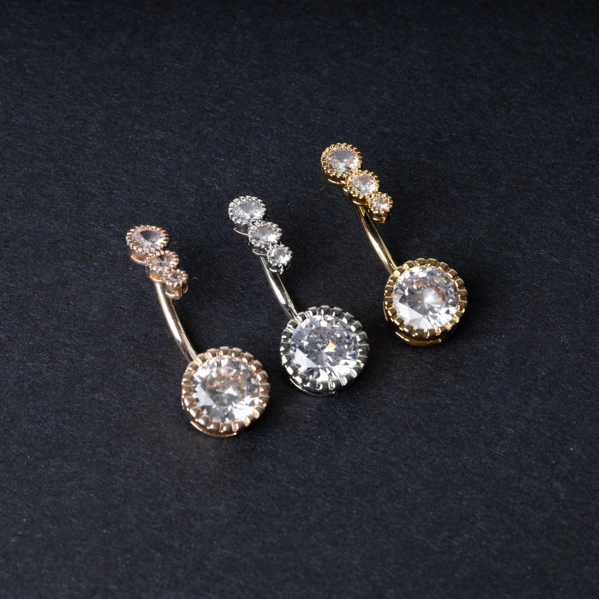Double-jeweled belly rings in gold, silver, and rose gold finishes with large and small crystal accents, displayed on a dark background.