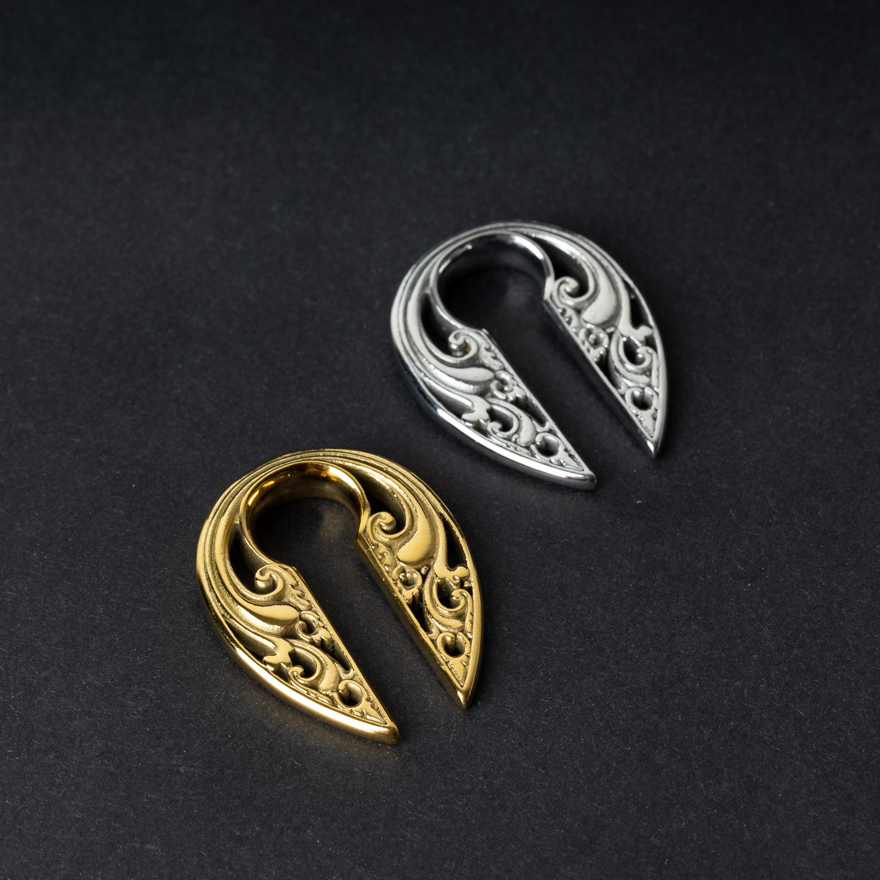 Two split ear weights with intricate filigree detailing, one in polished gold and the other in polished silver, displayed on a dark background.

