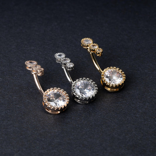 Double-jeweled belly rings in gold, silver, and rose gold finishes with large and small crystal accents, displayed on a dark background.