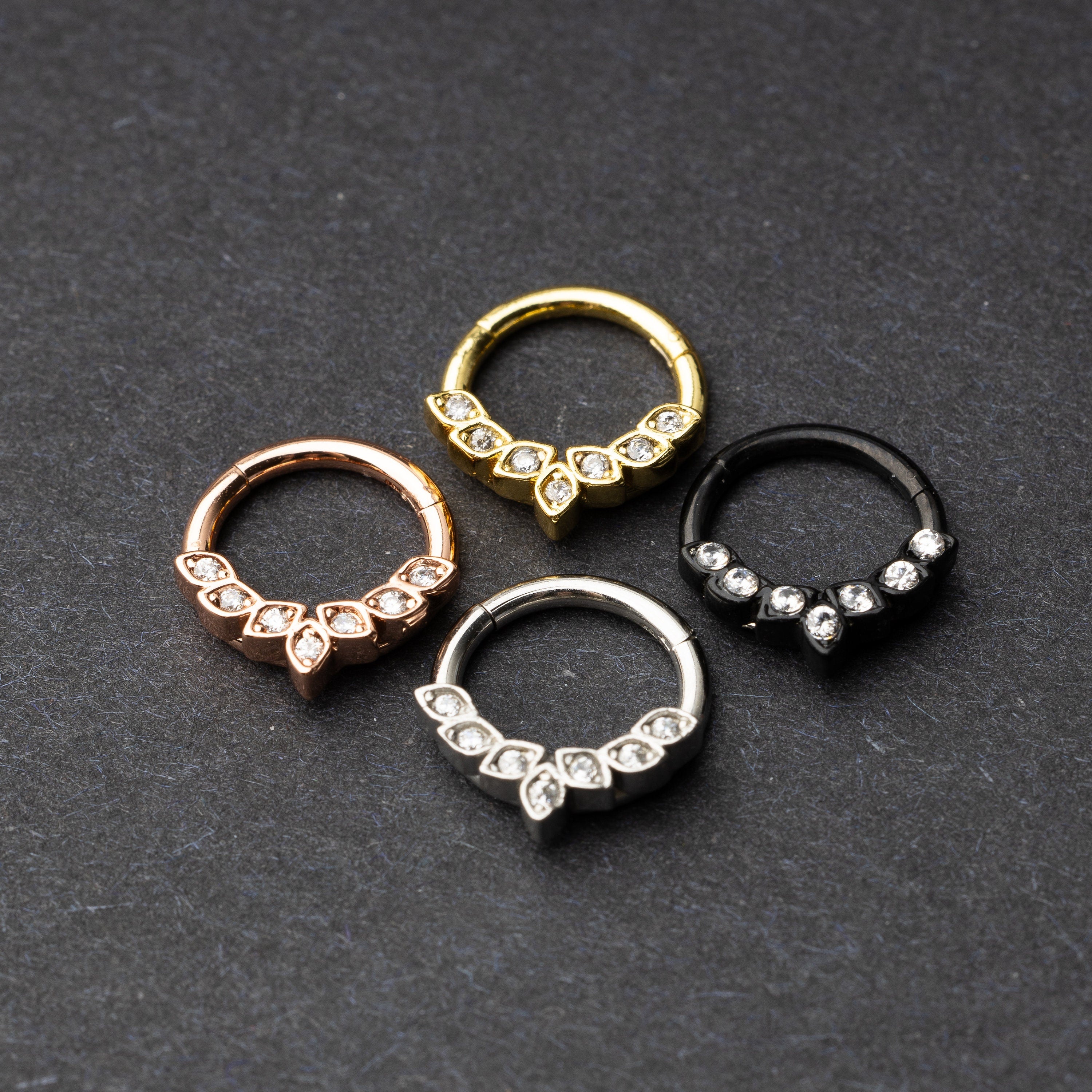 Four marquise gemstone stainless steel clickers in rose gold, gold, silver, and black finishes, arranged on a dark textured background.