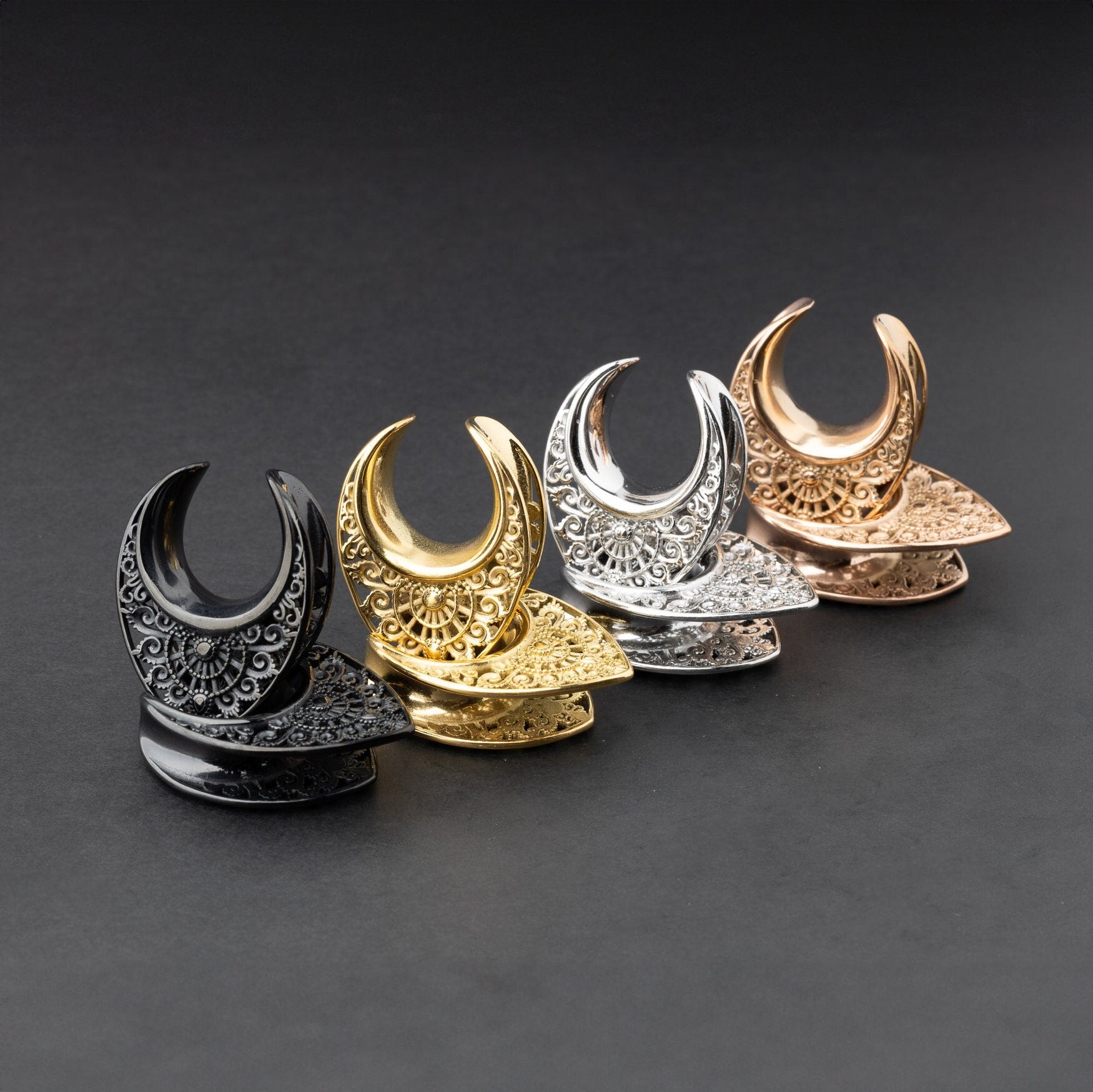 Crescent moon plugs for stretched ears in black, gold, silver, and rose gold finishes, displayed against a dark background.