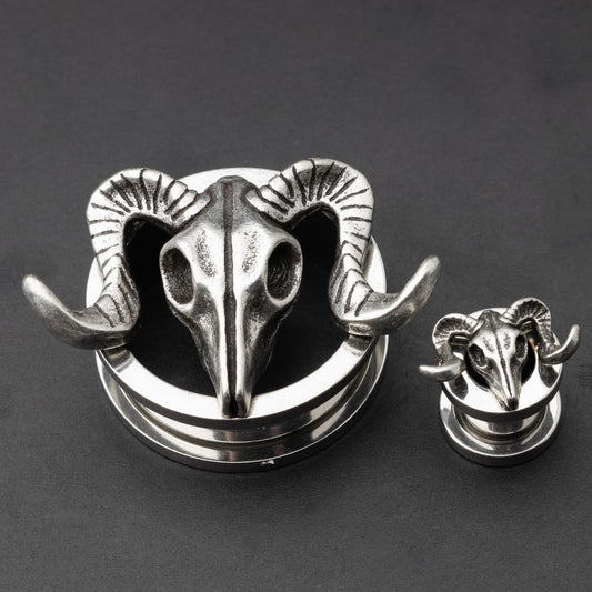 A pair of stainless steel saddle plugs featuring a 3D ram skull design, displayed on a dark, textured background.