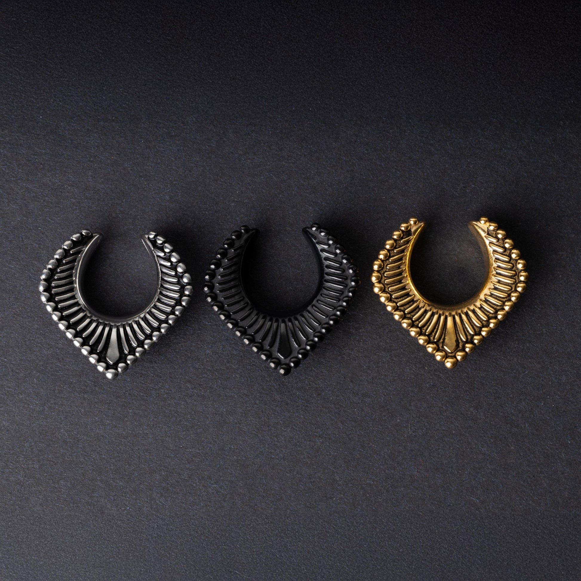 Three V-shaped stainless steel clickers in silver, black, and gold finishes arranged on a dark textured background.