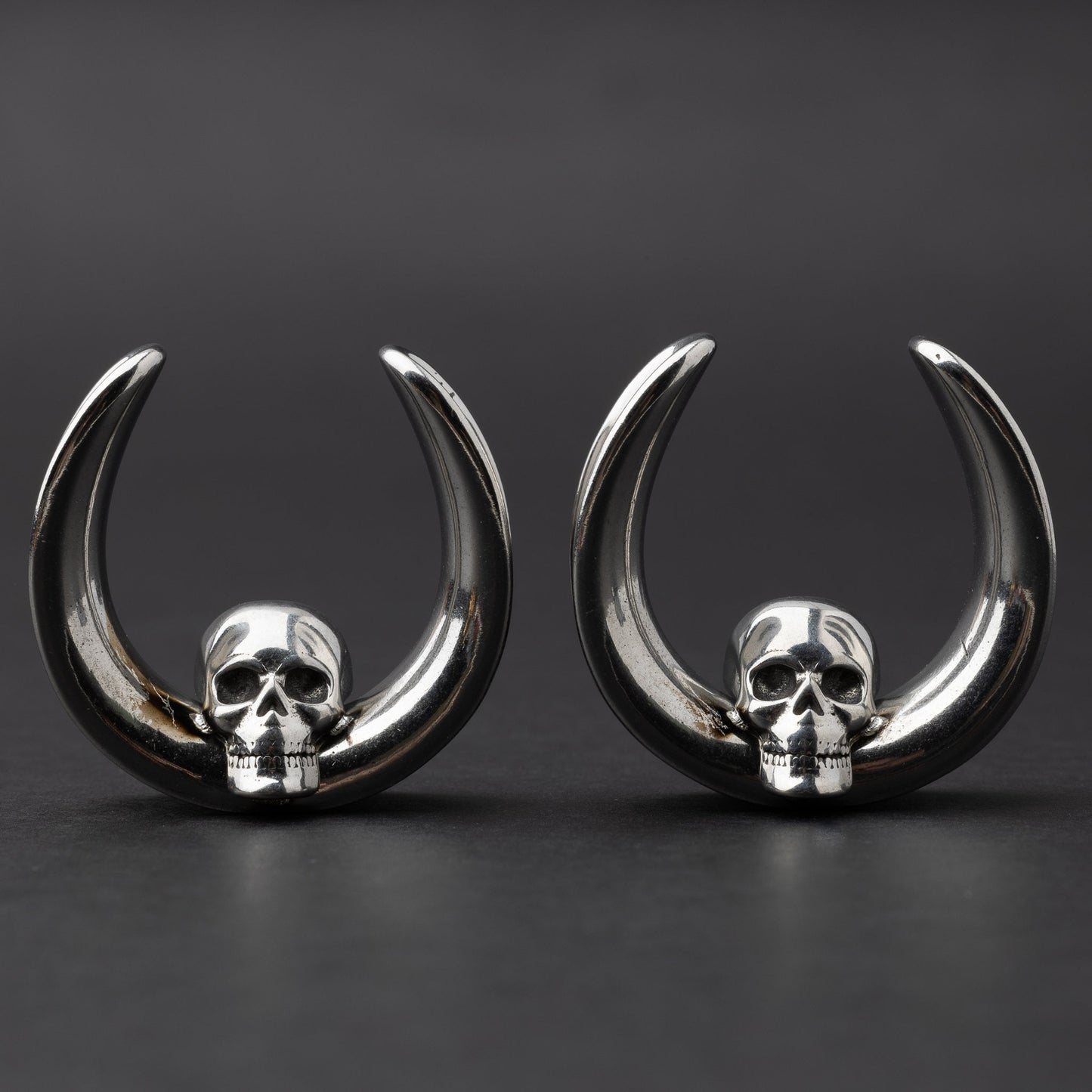 Pair of Skull design saddle tunnels in silver, displayed on a dark background.
