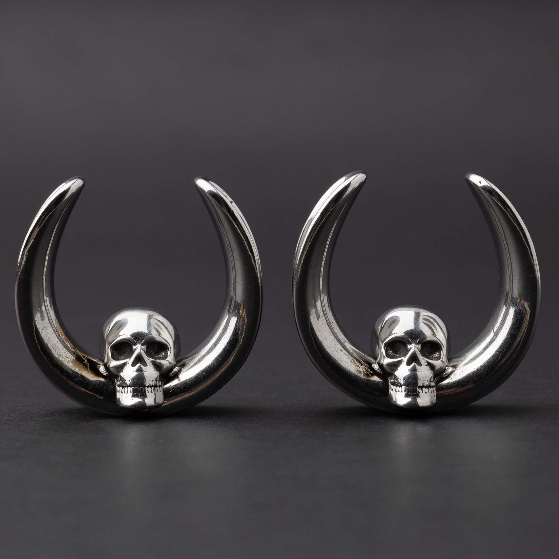 Pair of Skull design saddle tunnels in silver, displayed on a dark background.