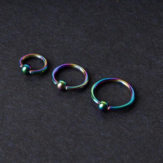 Three iridescent titanium captive bead rings of varying sizes displayed on a dark textured background, showcasing their vibrant rainbow finish.