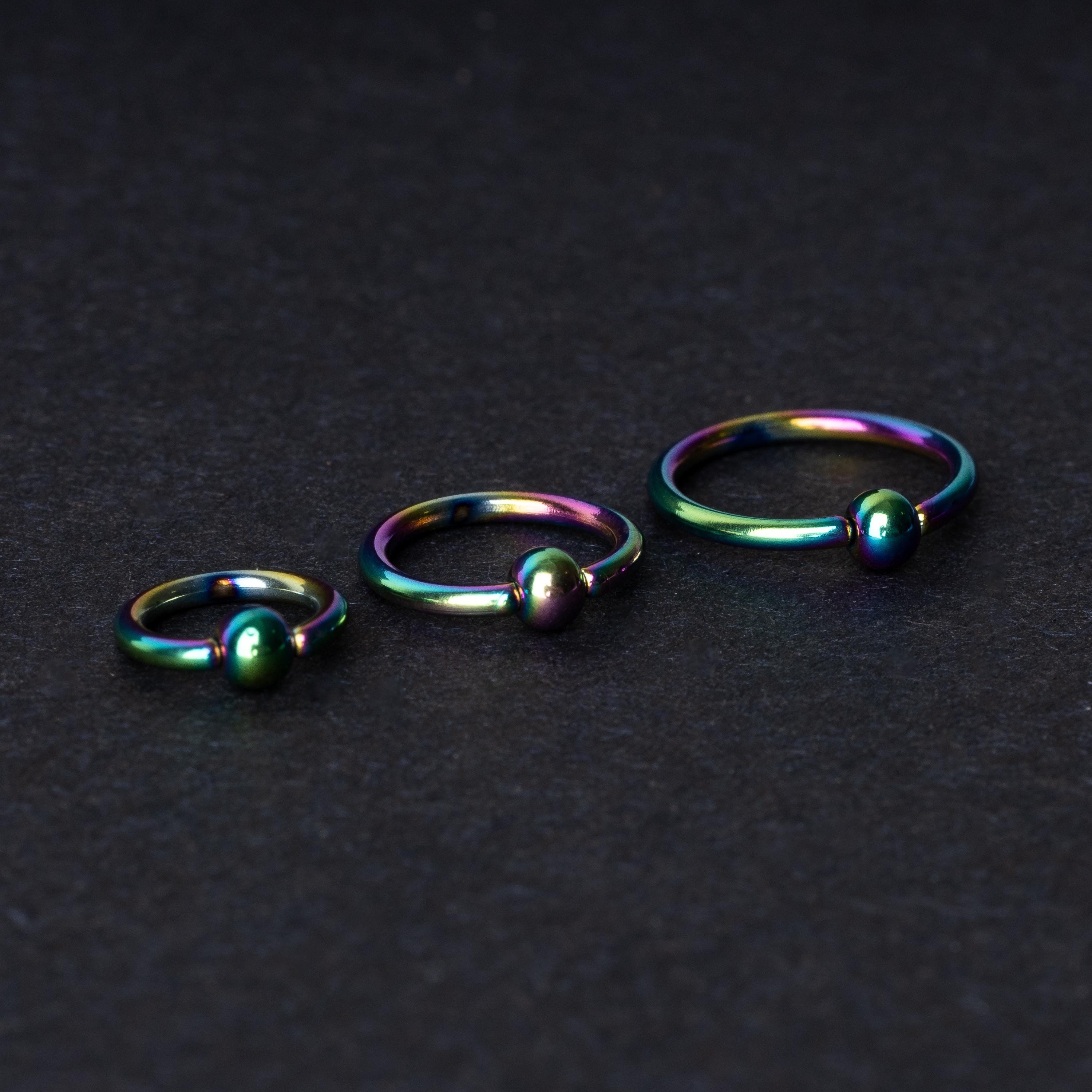 Three iridescent titanium captive bead rings of varying sizes displayed on a dark textured background, showcasing their vibrant rainbow finish.