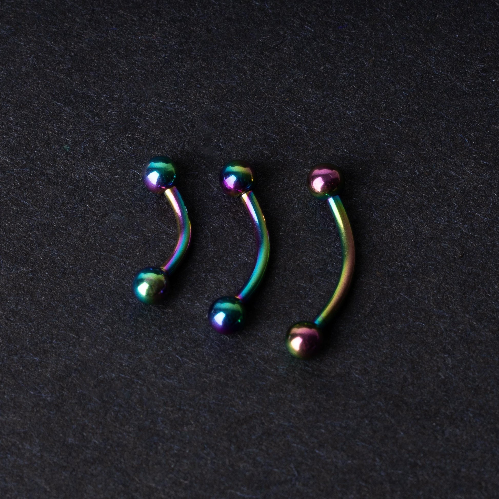 Three curved titanium barbells with a rainbow iridescent finish displayed on a dark textured background, highlighting their vivid colors and smooth finish.