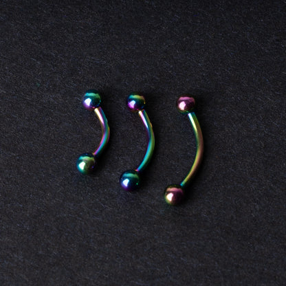 Three curved titanium barbells with a rainbow iridescent finish displayed on a dark textured background, highlighting their vivid colors and smooth finish.