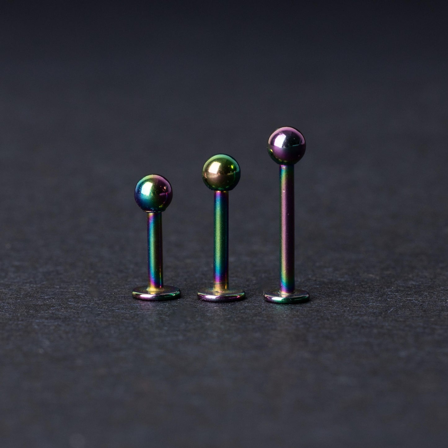 Three titanium labret studs with a rainbow iridescent finish, displayed upright on a dark textured background