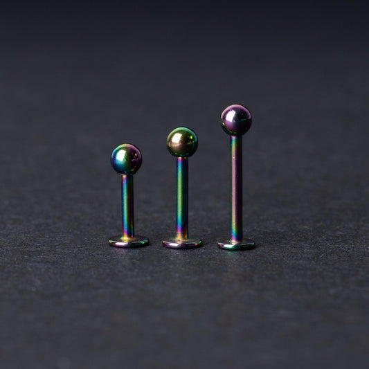 Three titanium labret studs with a rainbow iridescent finish, displayed upright on a dark textured background