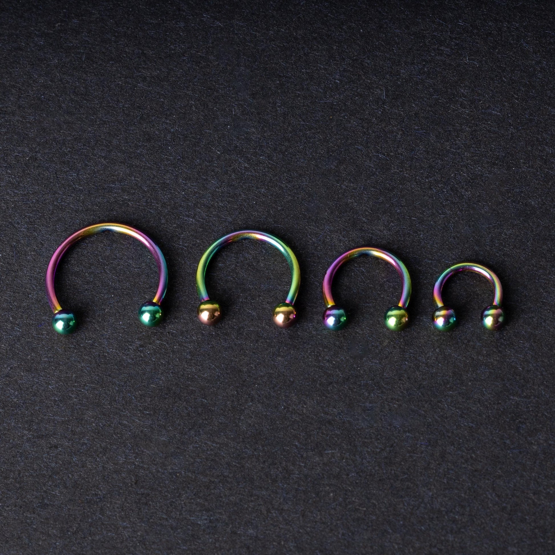 Close-up of four anodized rainbow horseshoe rings in varying sizes, with vibrant, multicolor finishes, displayed on a dark background.

