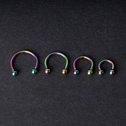 Close-up of four anodized rainbow horseshoe rings in varying sizes, with vibrant, multicolor finishes, displayed on a dark background.

