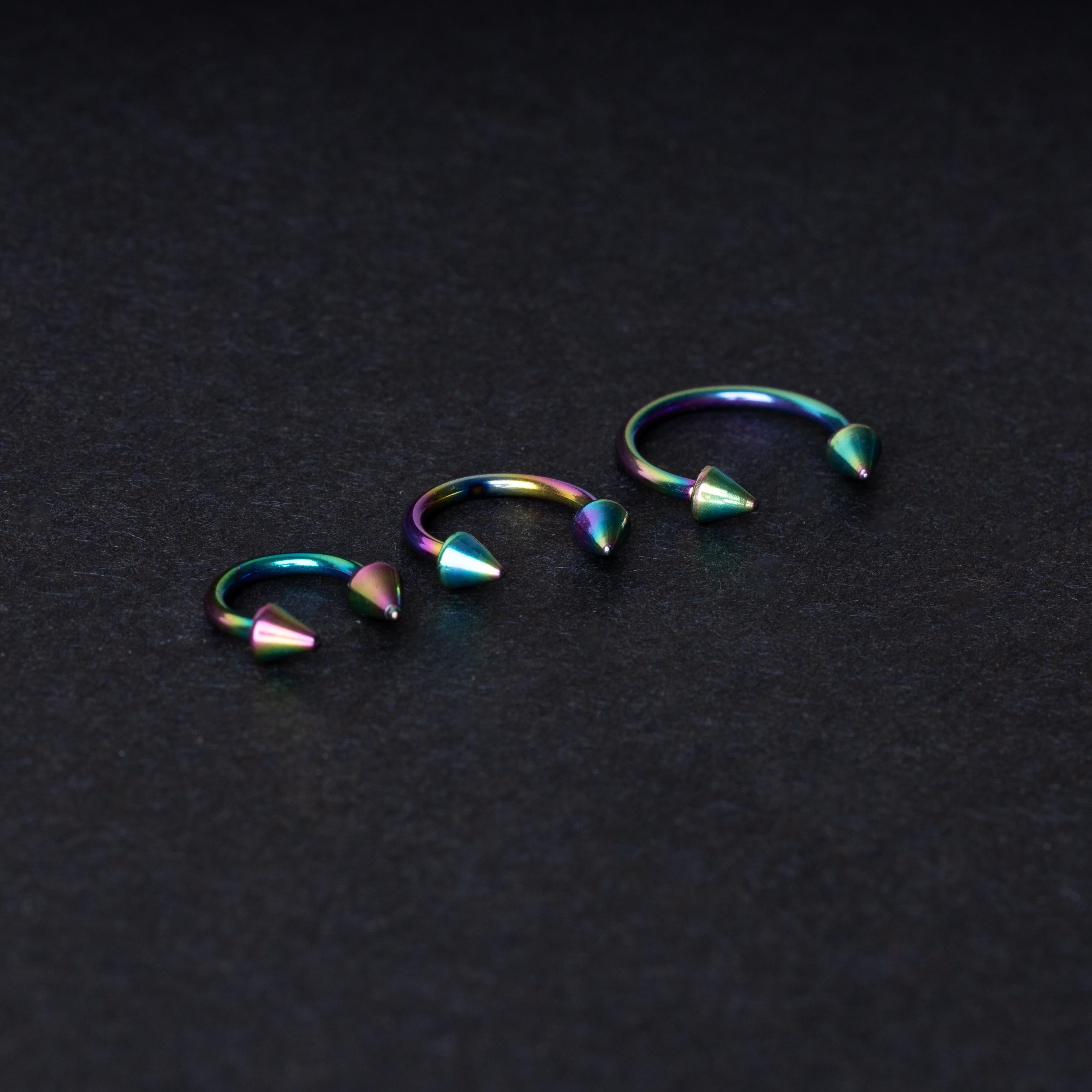 Three stainless steel horseshoe hoops with cone-shaped ends and a rainbow anodized finish displayed on a dark background, showcasing their bold design and vibrant colors.