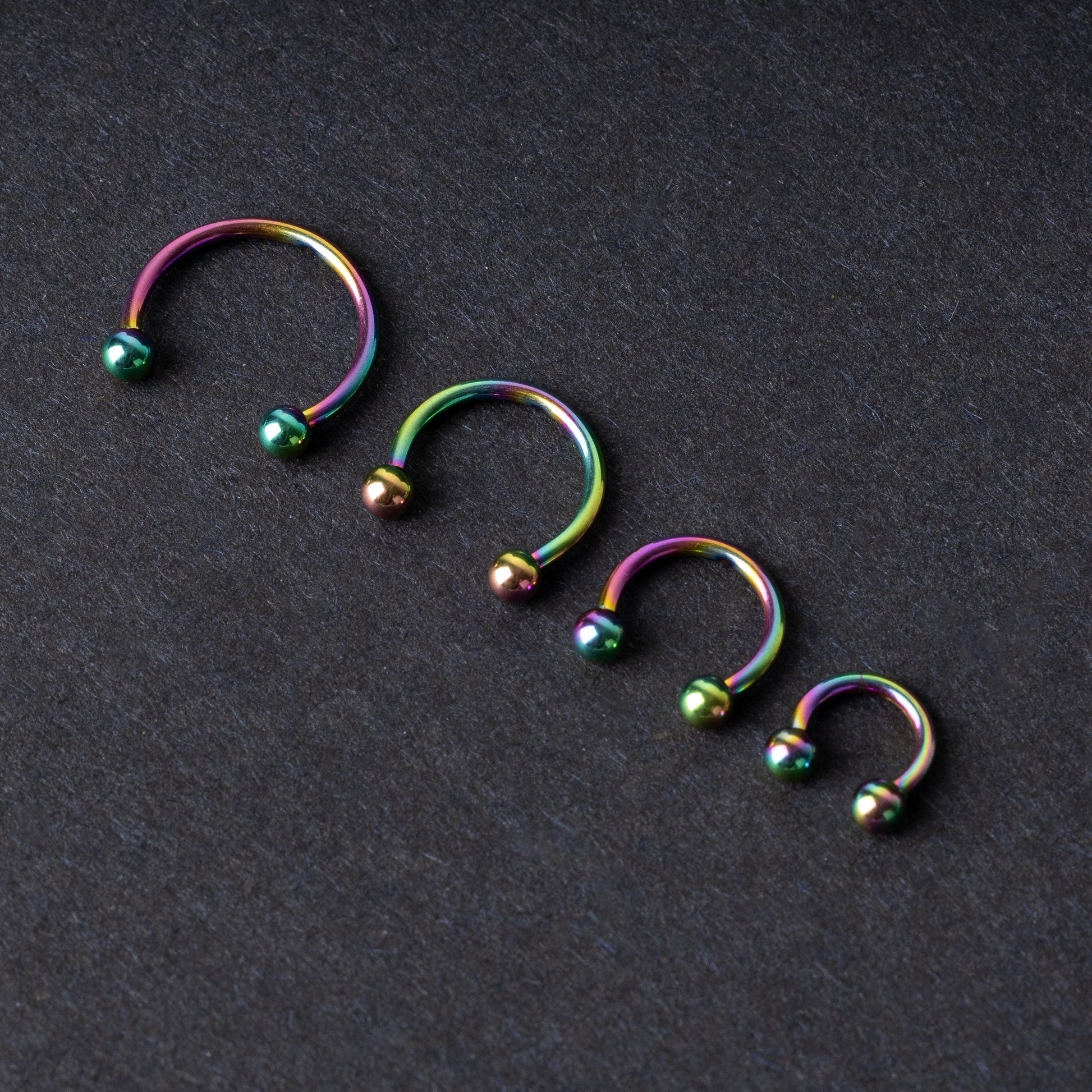 Close-up of four anodized rainbow horseshoe rings in varying sizes, with vibrant, multicolor finishes, displayed on a dark background.

