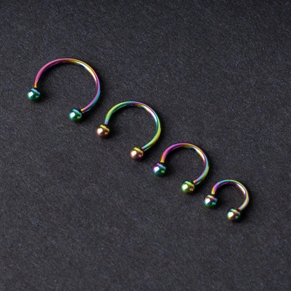 Close-up of four anodized rainbow horseshoe rings in varying sizes, with vibrant, multicolor finishes, displayed on a dark background.

