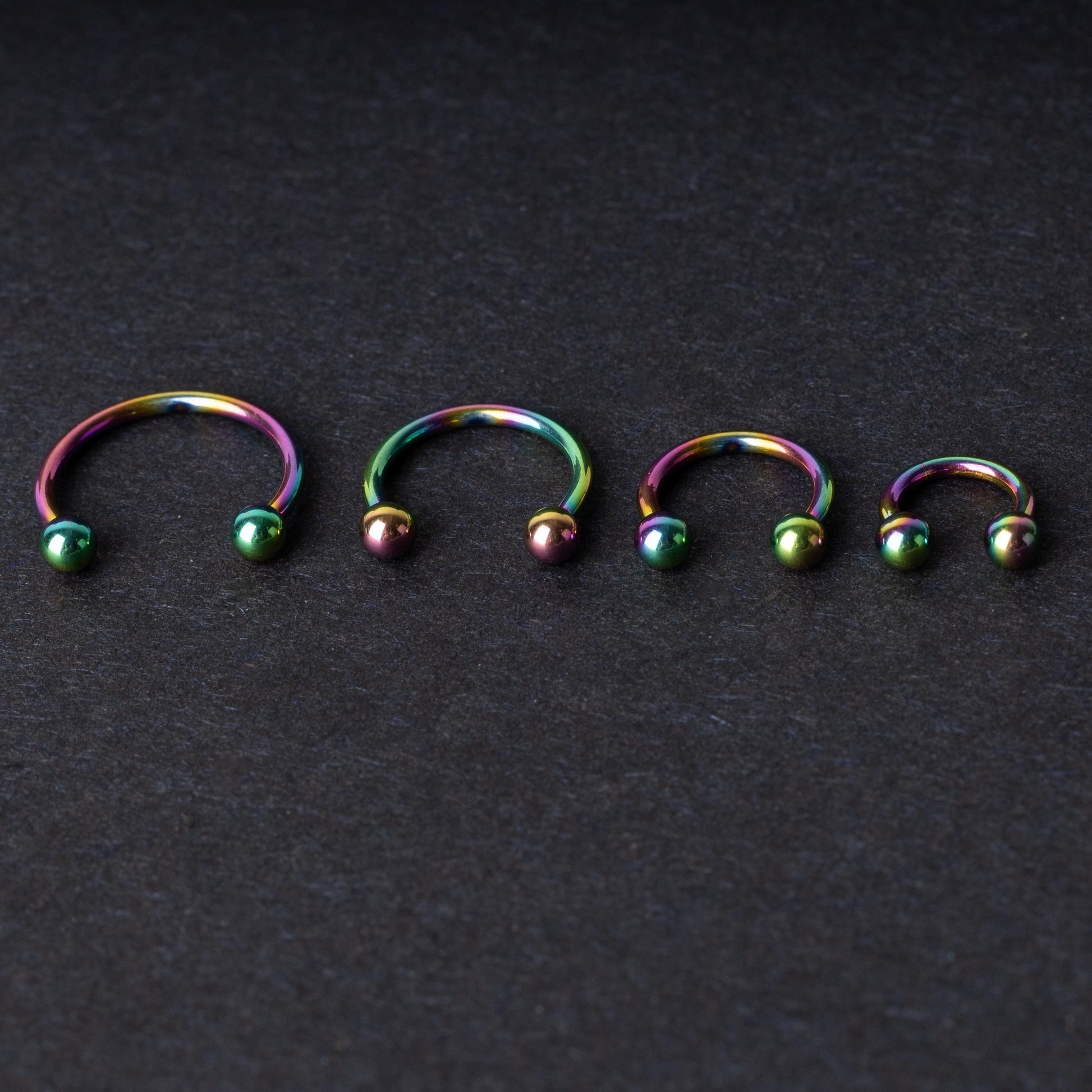 Close-up of four anodized rainbow horseshoe rings in varying sizes, with vibrant, multicolor finishes, displayed on a dark background.

