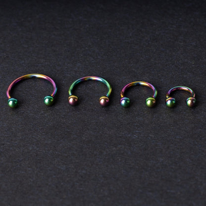 Close-up of four anodized rainbow horseshoe rings in varying sizes, with vibrant, multicolor finishes, displayed on a dark background.

