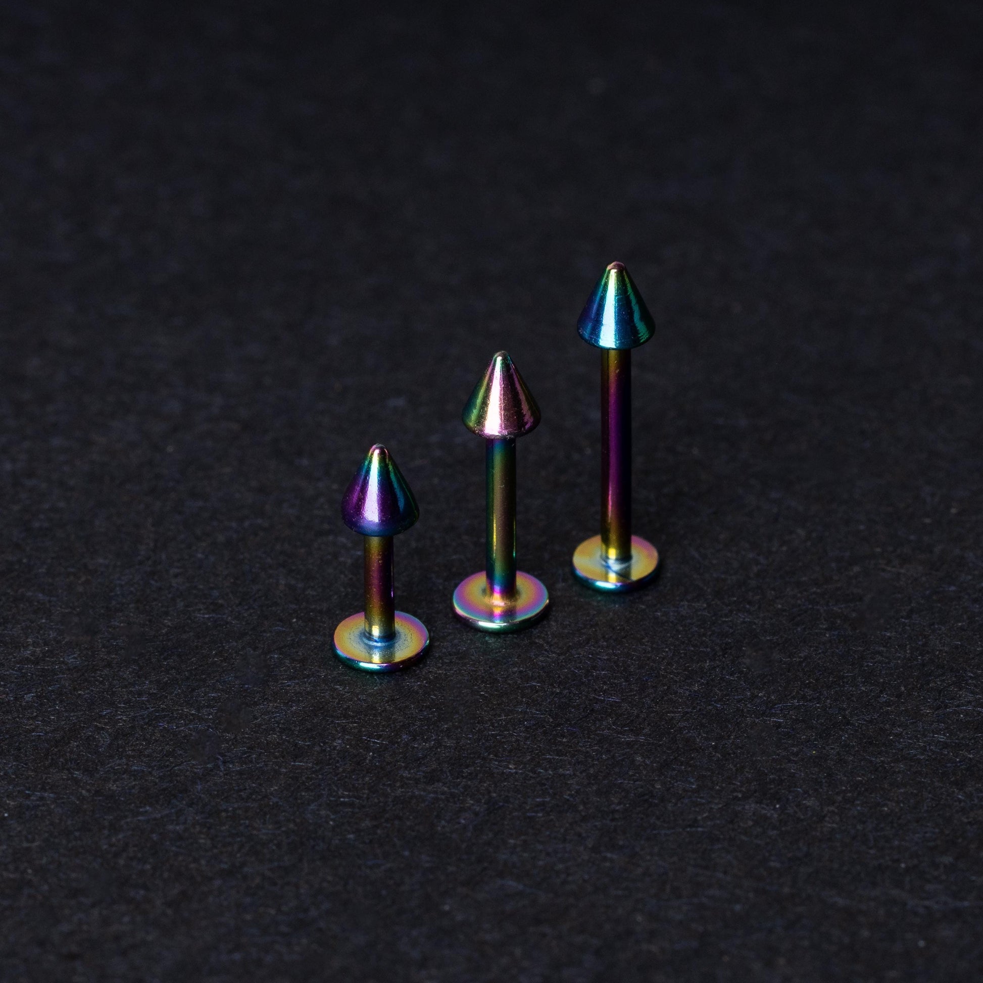 Three rainbow labret studs with cone-shaped tops displayed on a dark background, highlighting their iridescent finish and sleek, durable design.