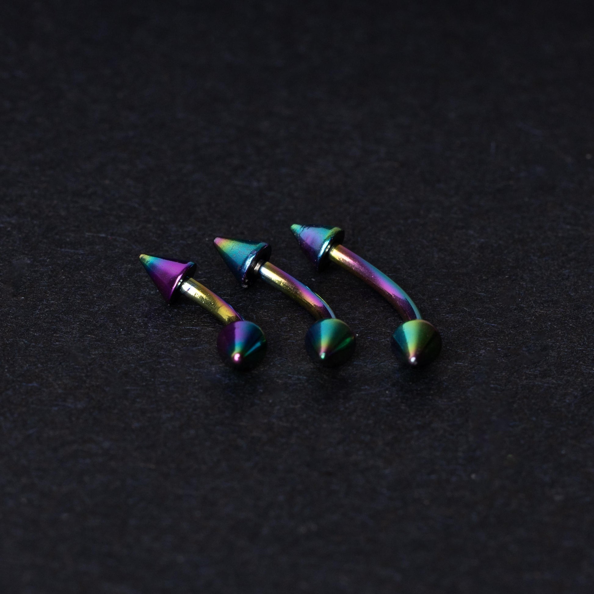 Three rainbow titanium curved barbells with cone-shaped ends, displayed on a dark textured background, showcasing their vivid color gradient and sleek design.