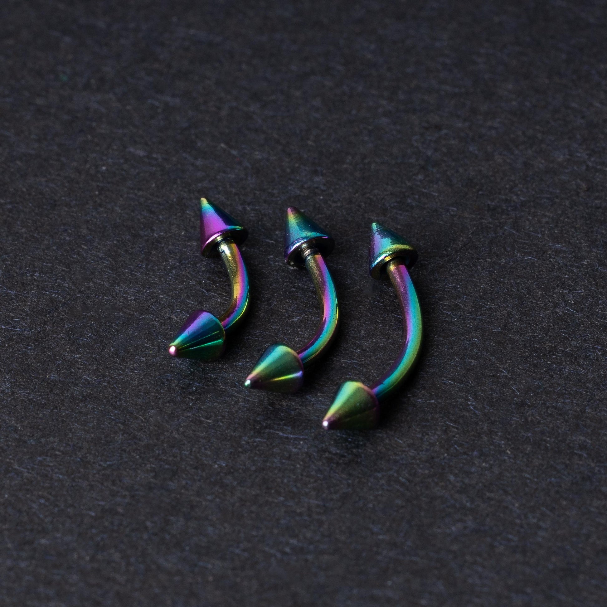 Three rainbow titanium curved barbells with cone-shaped ends, displayed on a dark textured background, showcasing their vivid color gradient and sleek design.