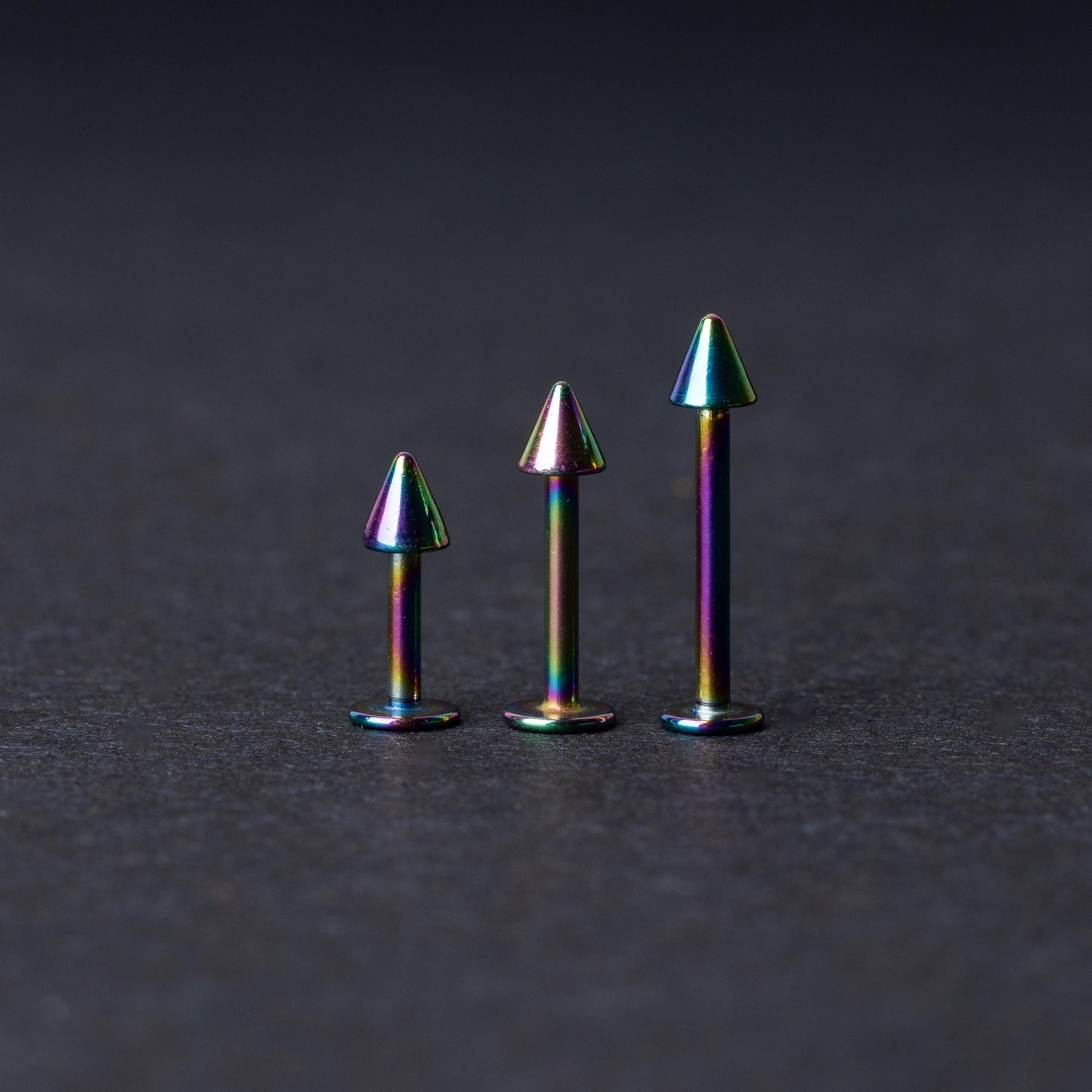 Three rainbow labret studs with cone-shaped tops displayed on a dark background, highlighting their iridescent finish and sleek, durable design.