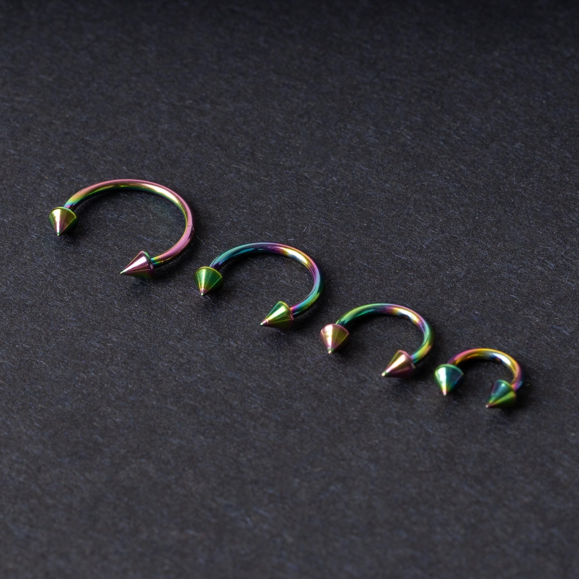 Titanium horseshoe hoops with spike ends, featuring a colorful rainbow finish, displayed on a dark textured background.
