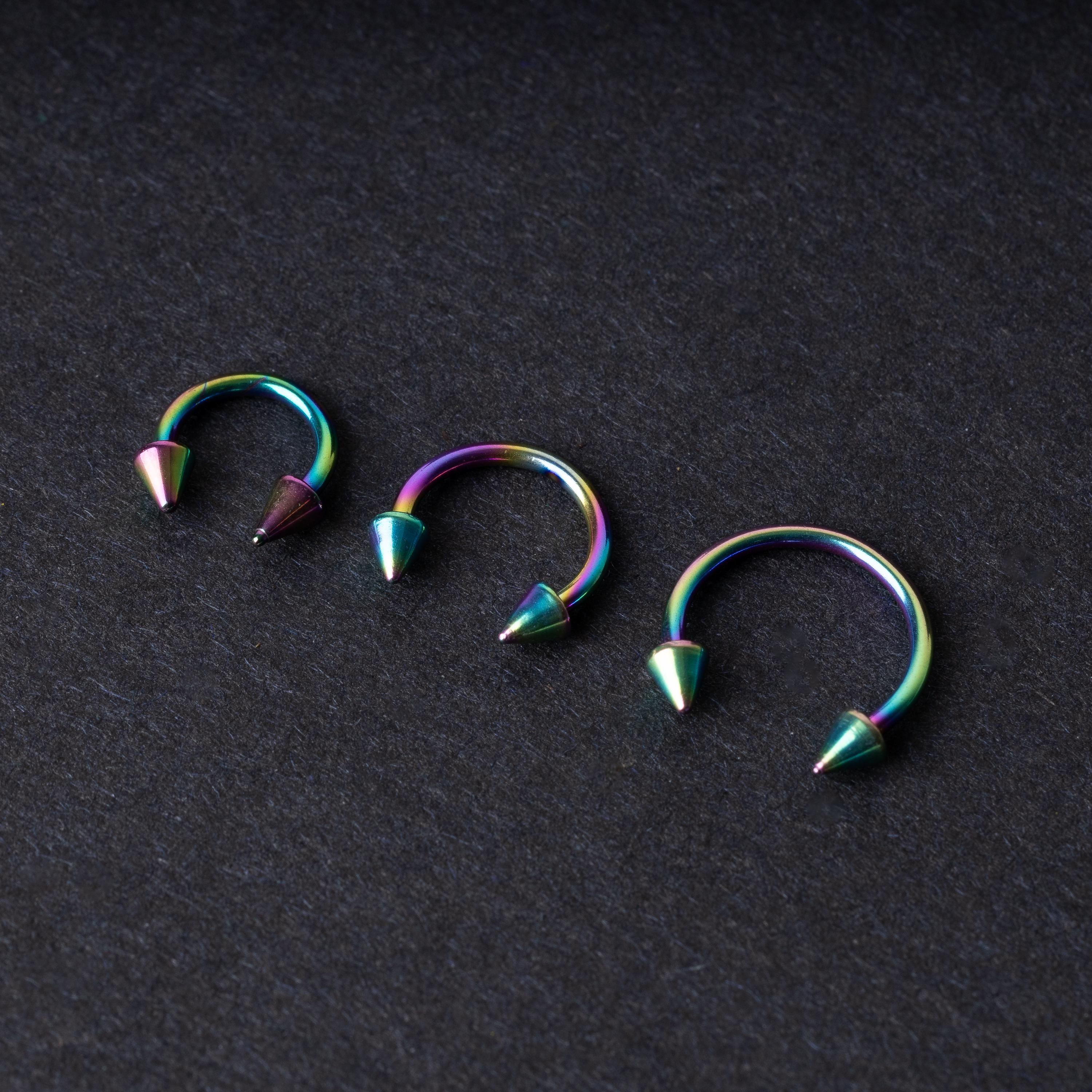 Three stainless steel horseshoe hoops with cone-shaped ends and a rainbow anodized finish displayed on a dark background, showcasing their bold design and vibrant colors.