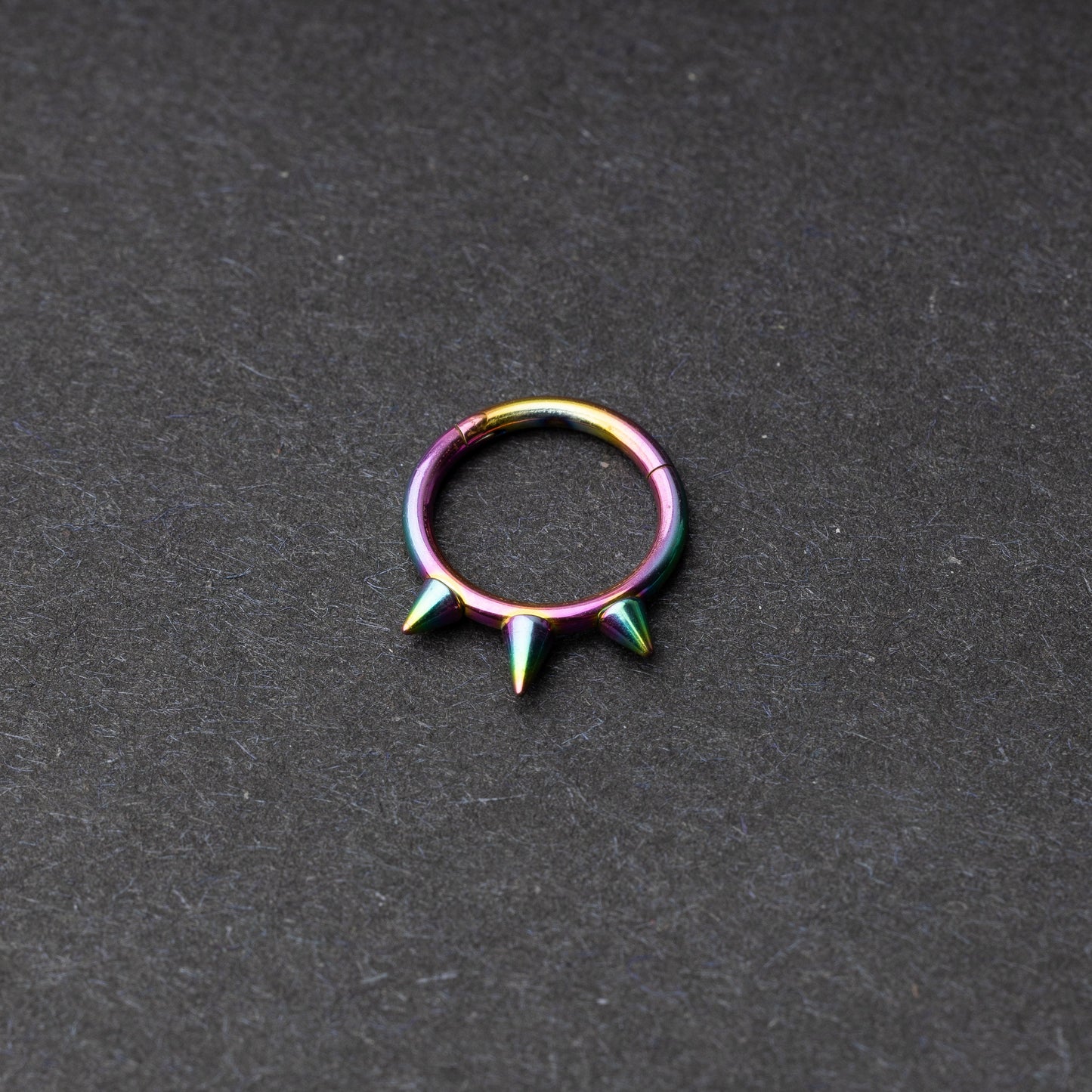 Rainbow-tone stainless steel clicker ring with three spikes, displayed on a dark textured background.