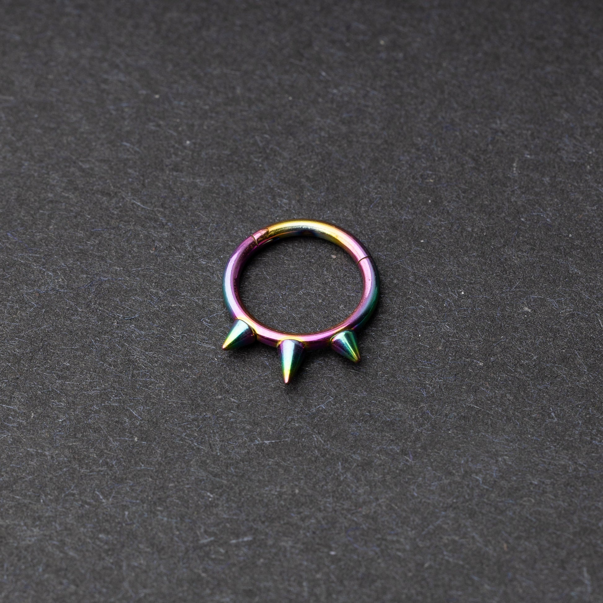 Rainbow-tone stainless steel clicker ring with three spikes, displayed on a dark textured background.