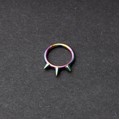 Rainbow-tone stainless steel clicker ring with three spikes, displayed on a dark textured background.