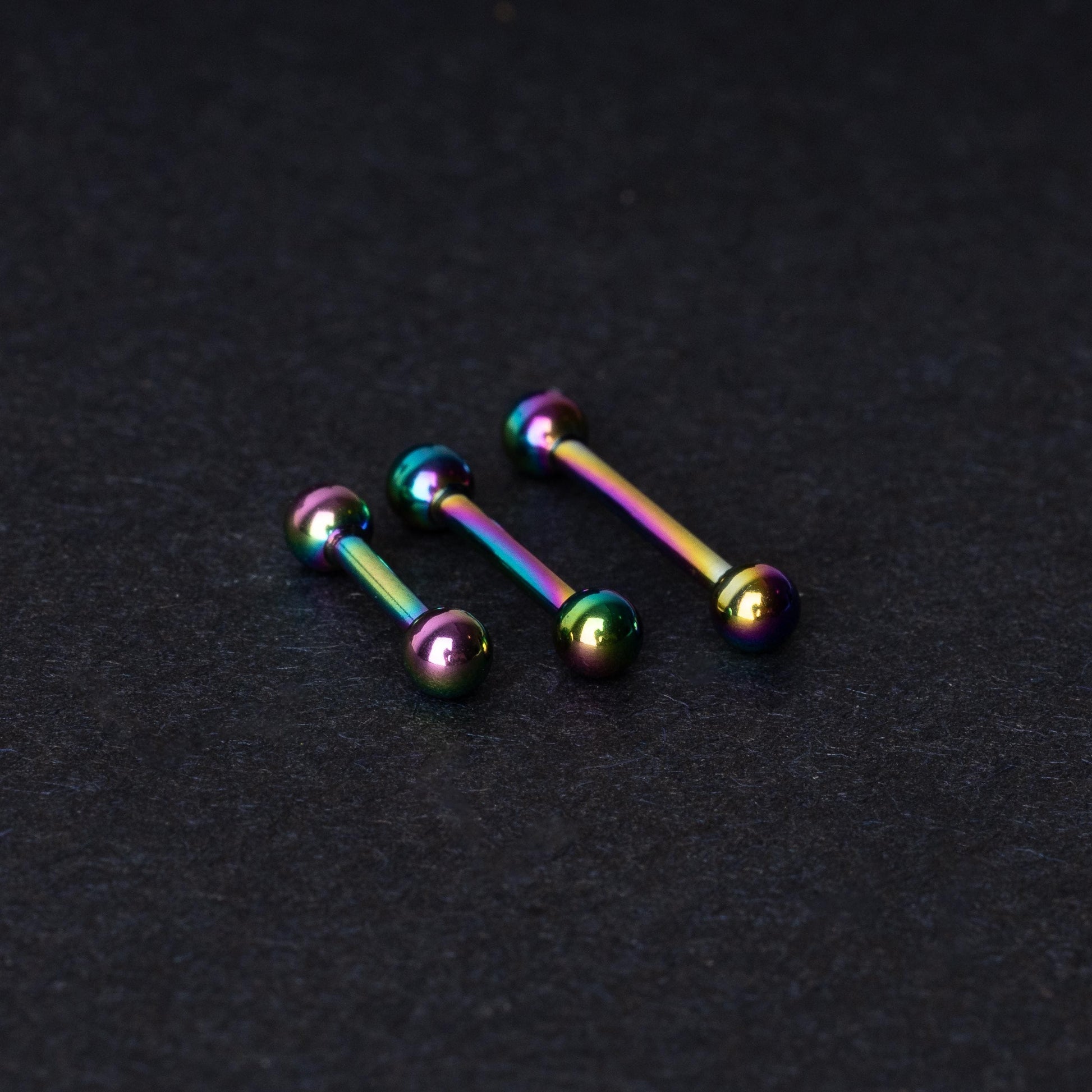 Rainbow stainless steel barbells with an anodized finish on a dark background.