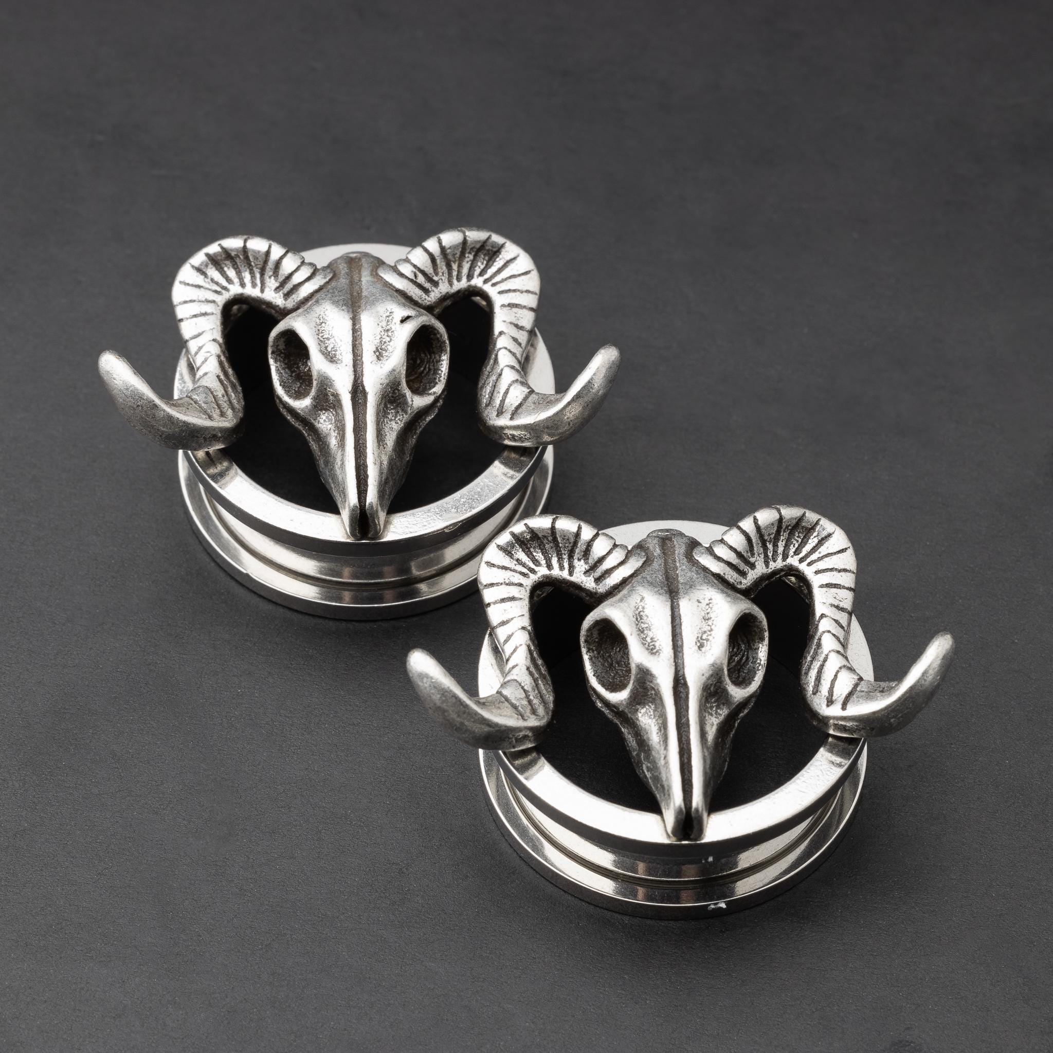 A pair of stainless steel saddle plugs featuring a 3D ram skull design, displayed on a dark, textured background.