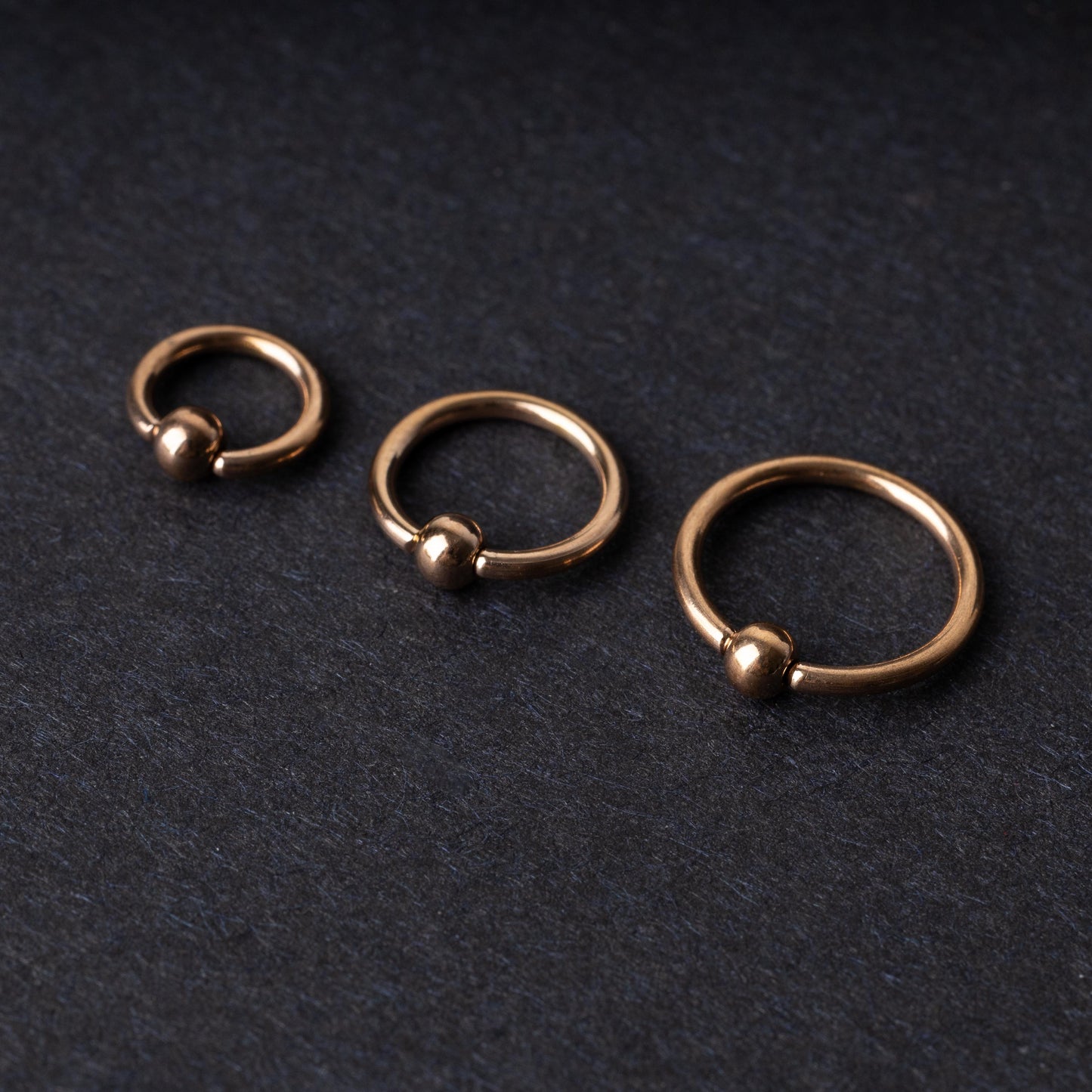 Rose Gold-tone ball closure rings in three sizes, displayed on a dark background.