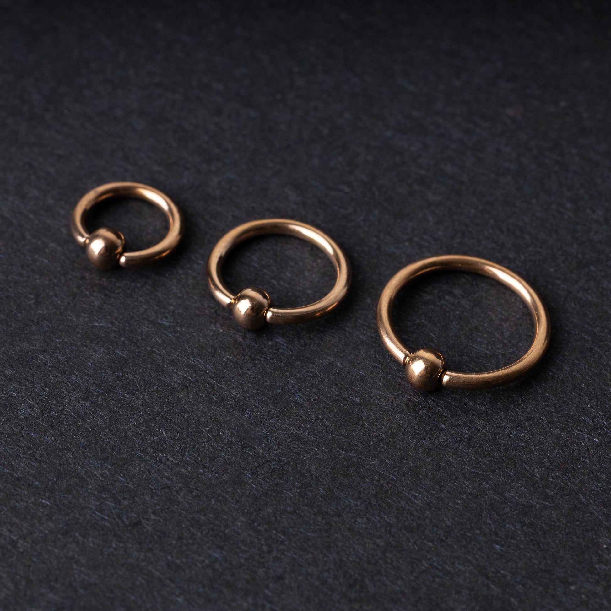Rose Gold-tone ball closure rings in three sizes, displayed on a dark background.